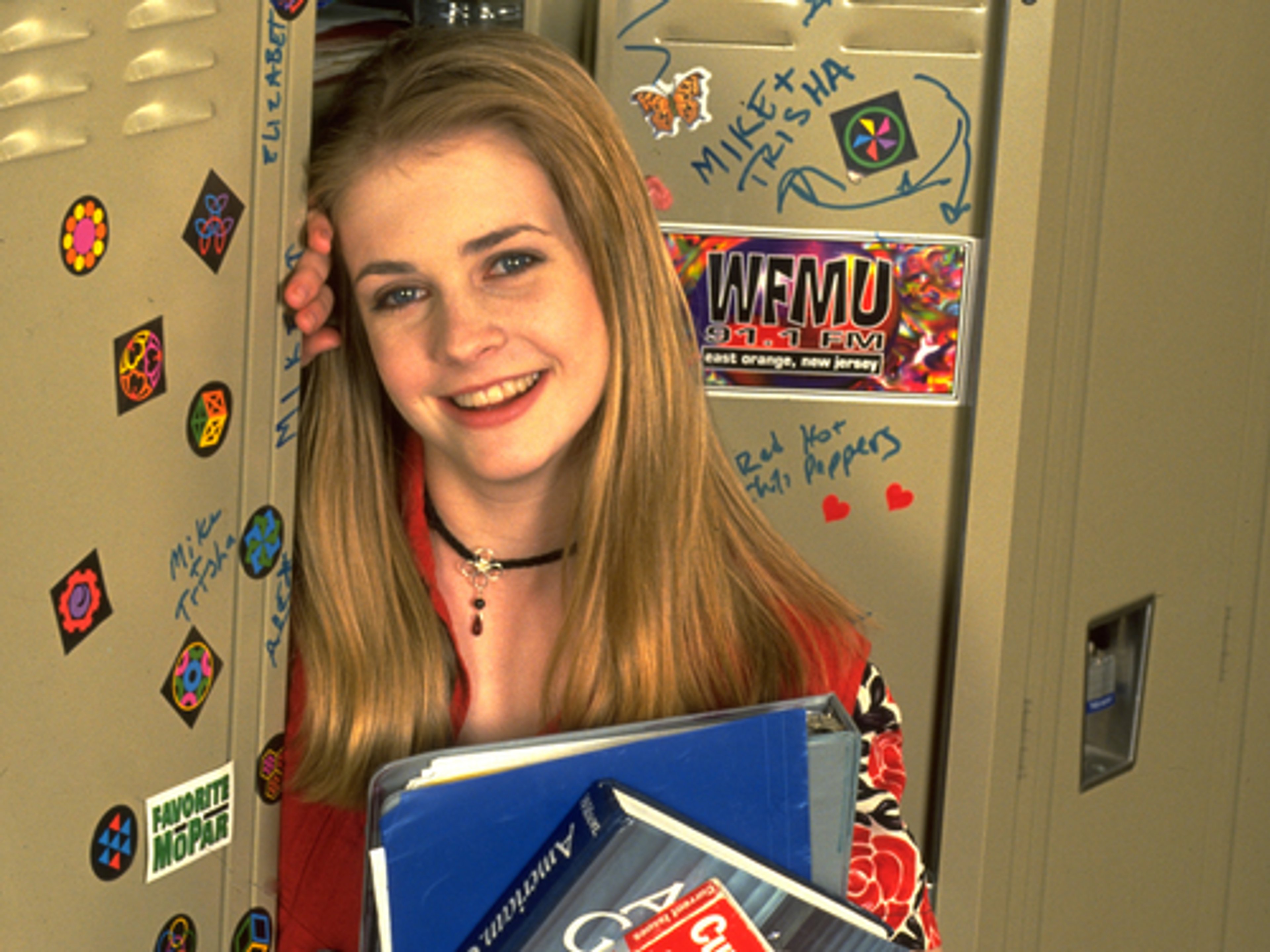 A young woman with long blonde hair, wearing a colorful, patterned shirt and holding a blue notebook, smiling at the camera.