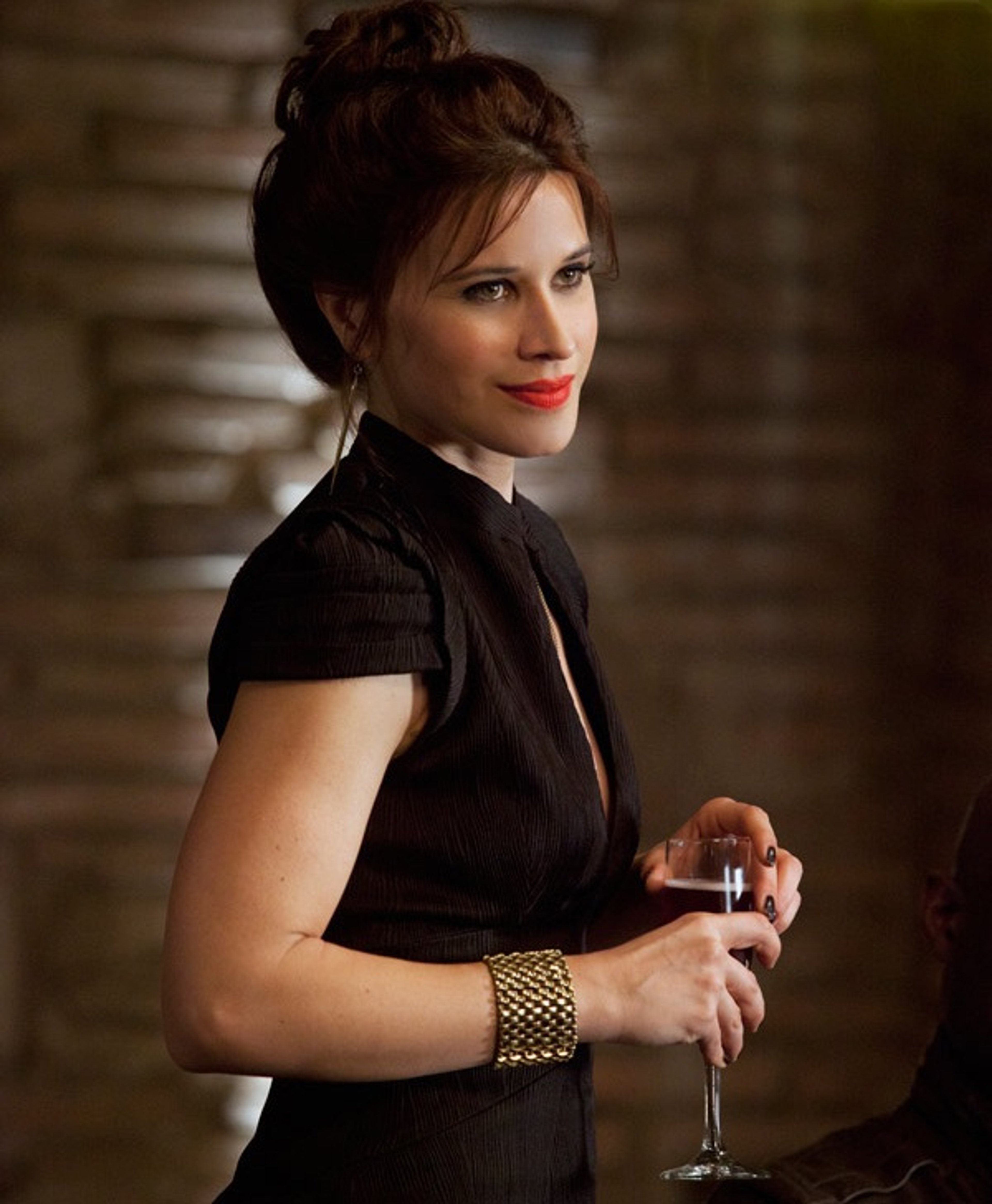 A woman with dark hair wearing a black dress, holding a glass of alcohol and looking serious.
