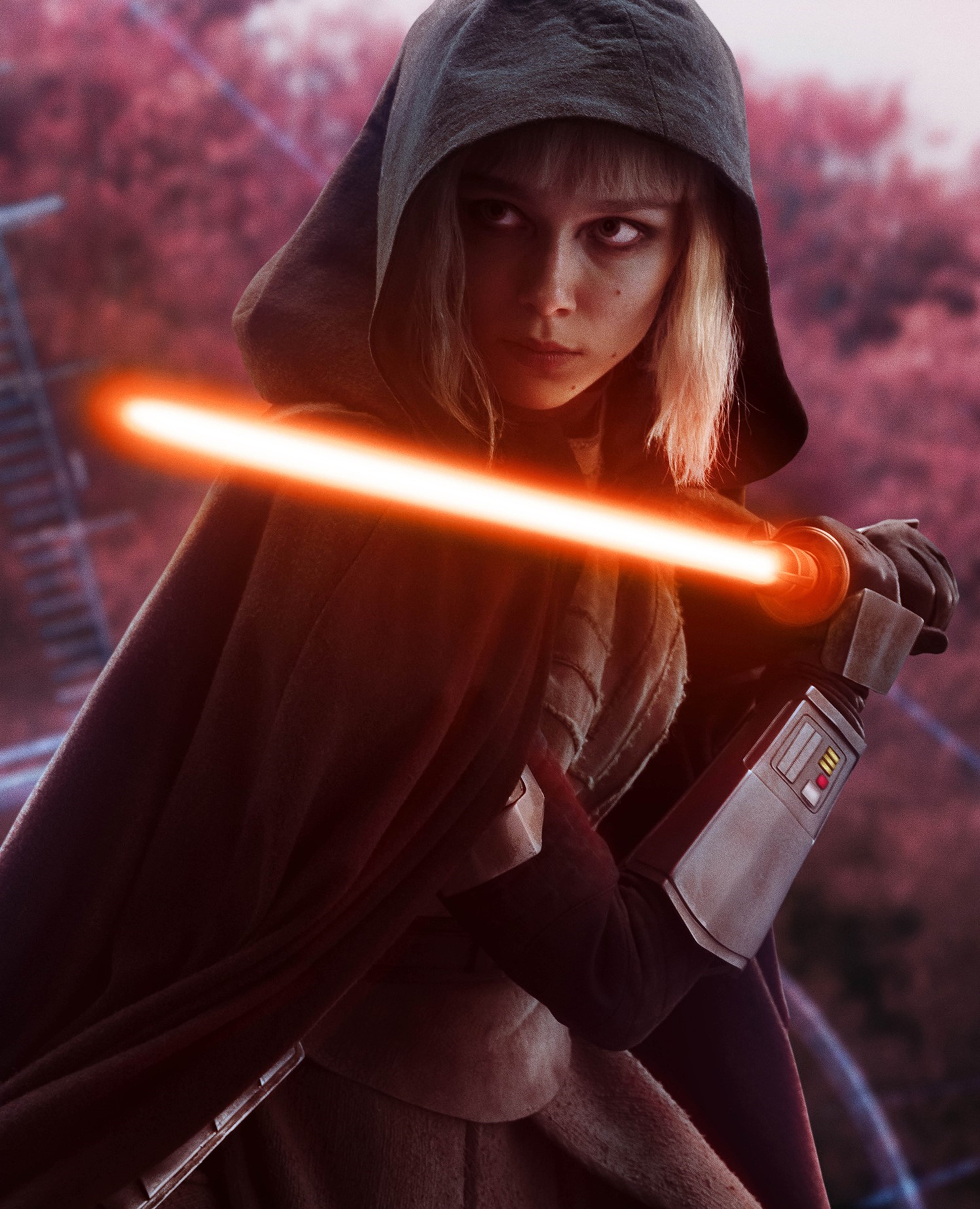 A woman in a dark hooded cloak holding a glowing orange lightsaber in a forest setting