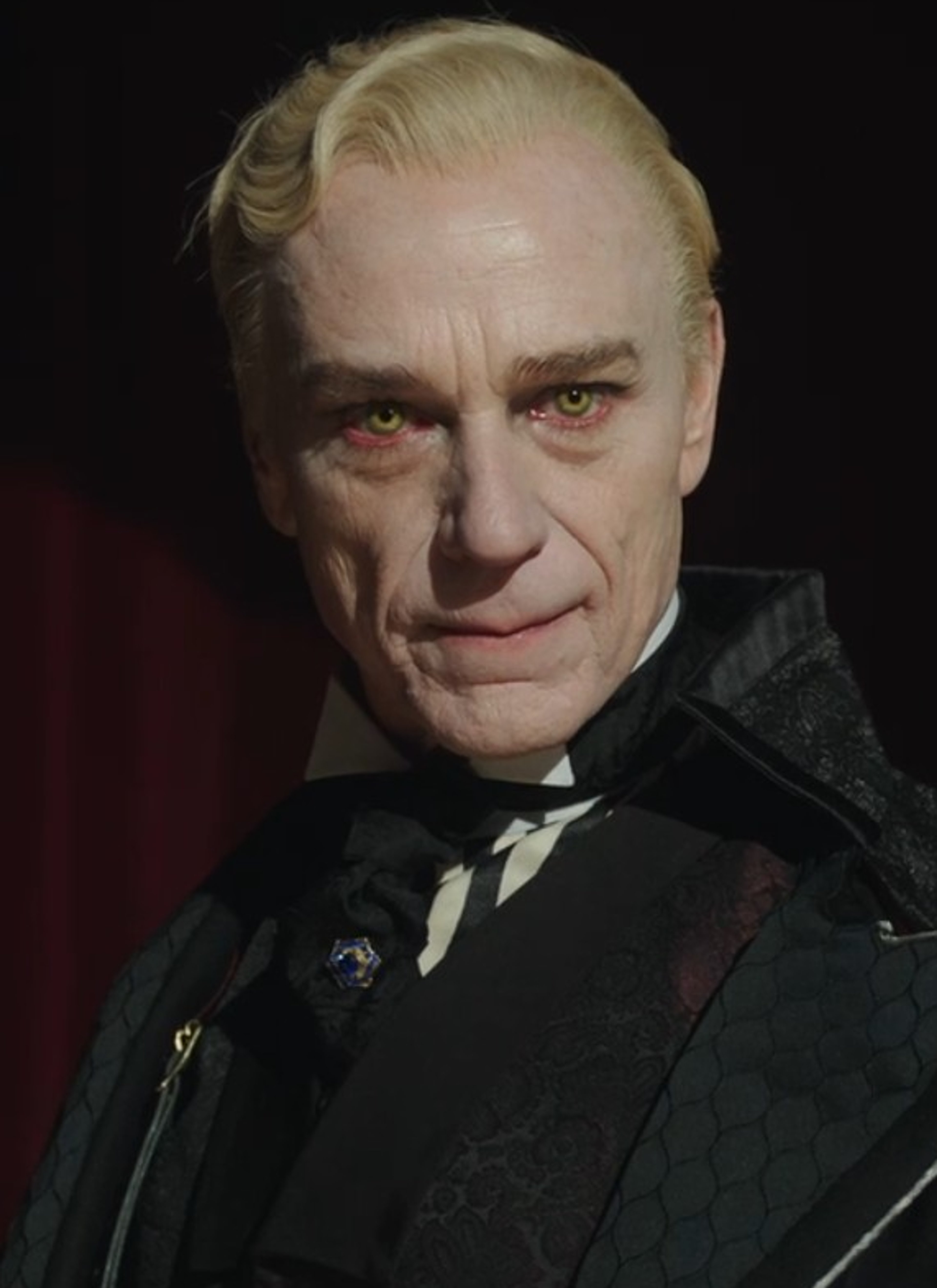 An older man with pale skin, white hair, and a stern expression wearing a black suit and tie