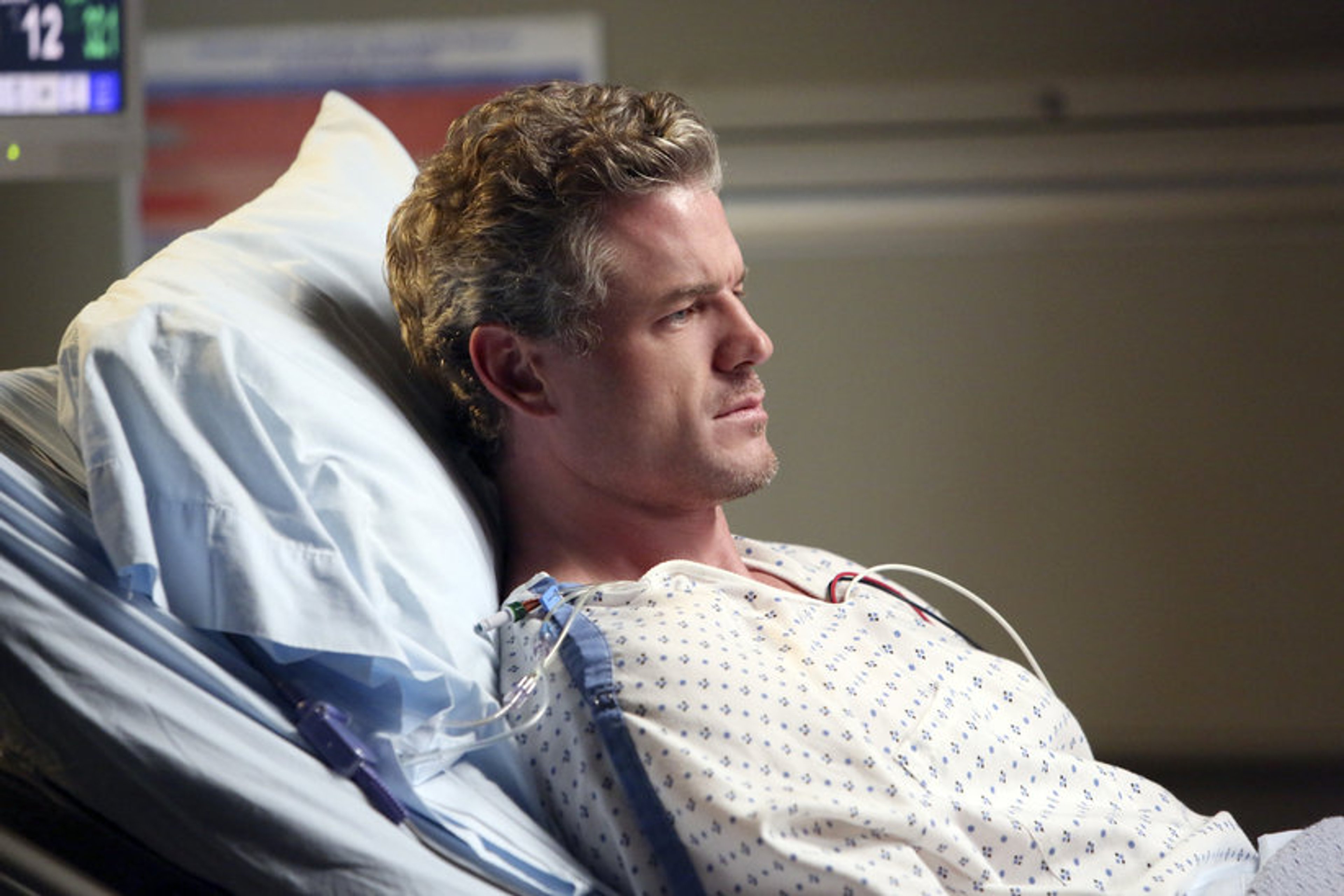 A middle-aged man with short brown hair and facial stubble lying in a hospital bed, wearing a hospital gown and with an IV line attached to his arm.
