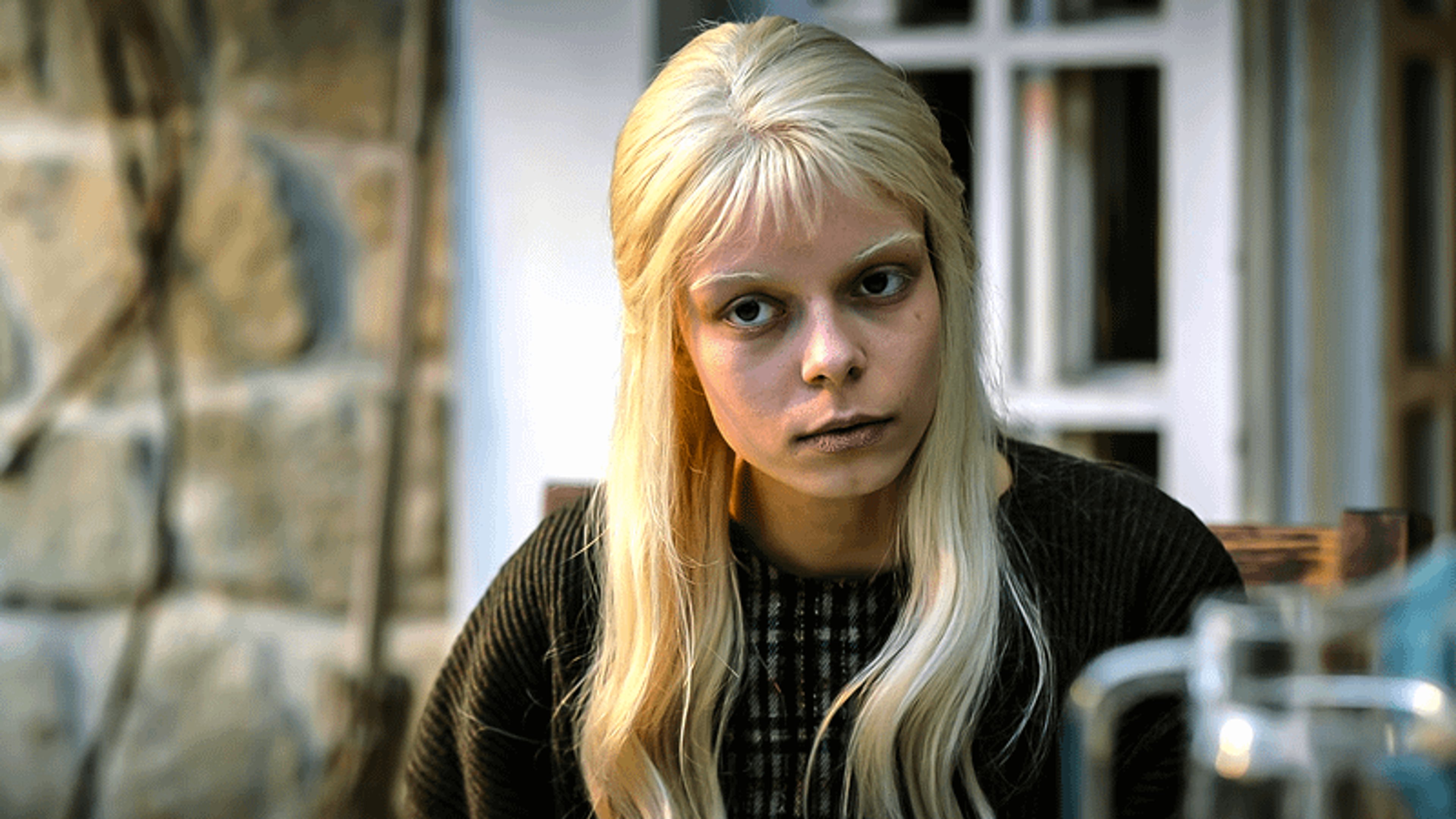 A young woman with blonde hair and a serious expression, wearing a black jacket.