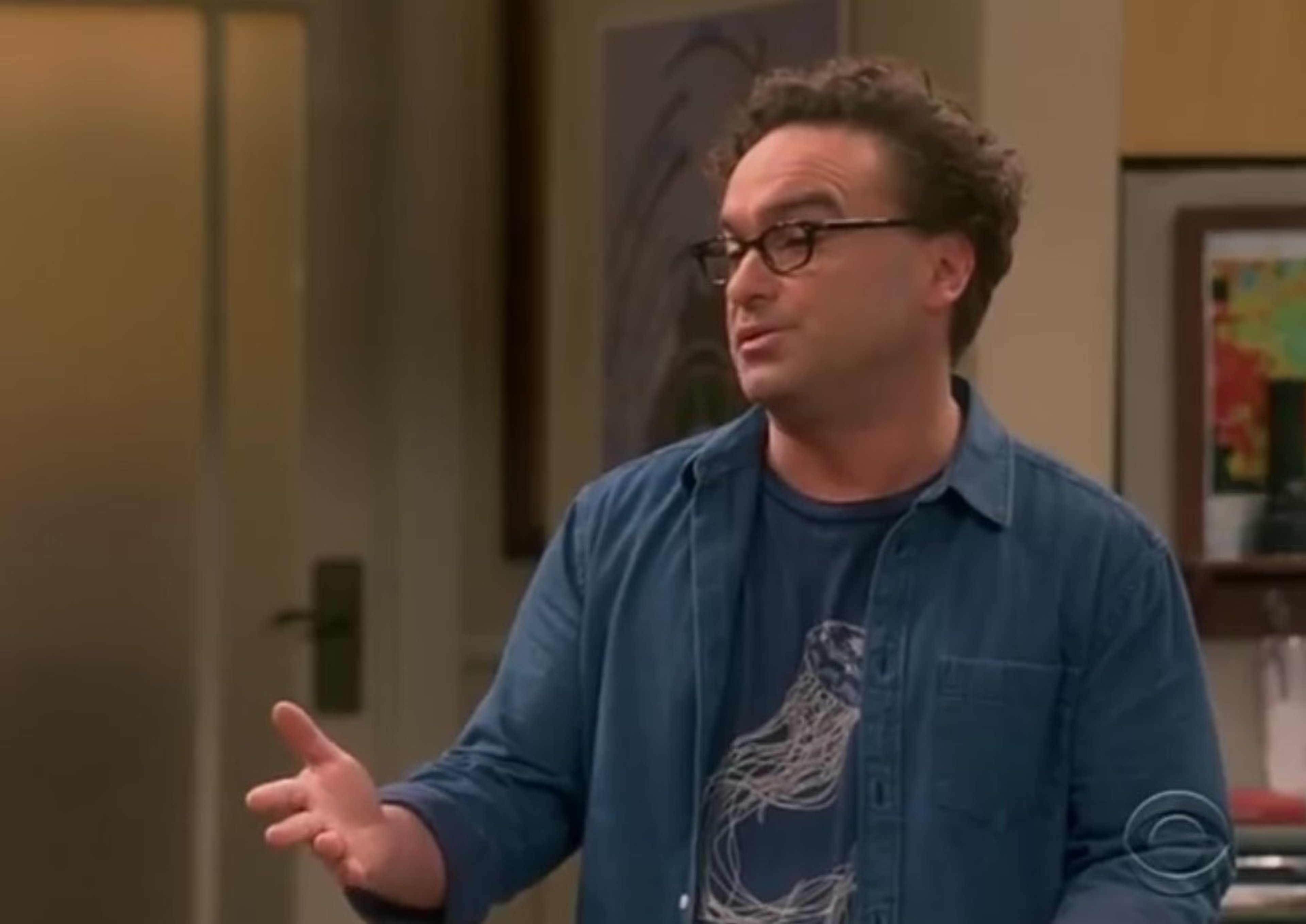 A middle-aged man with curly brown hair and glasses, wearing a blue button-down shirt, gesturing with his hands.
