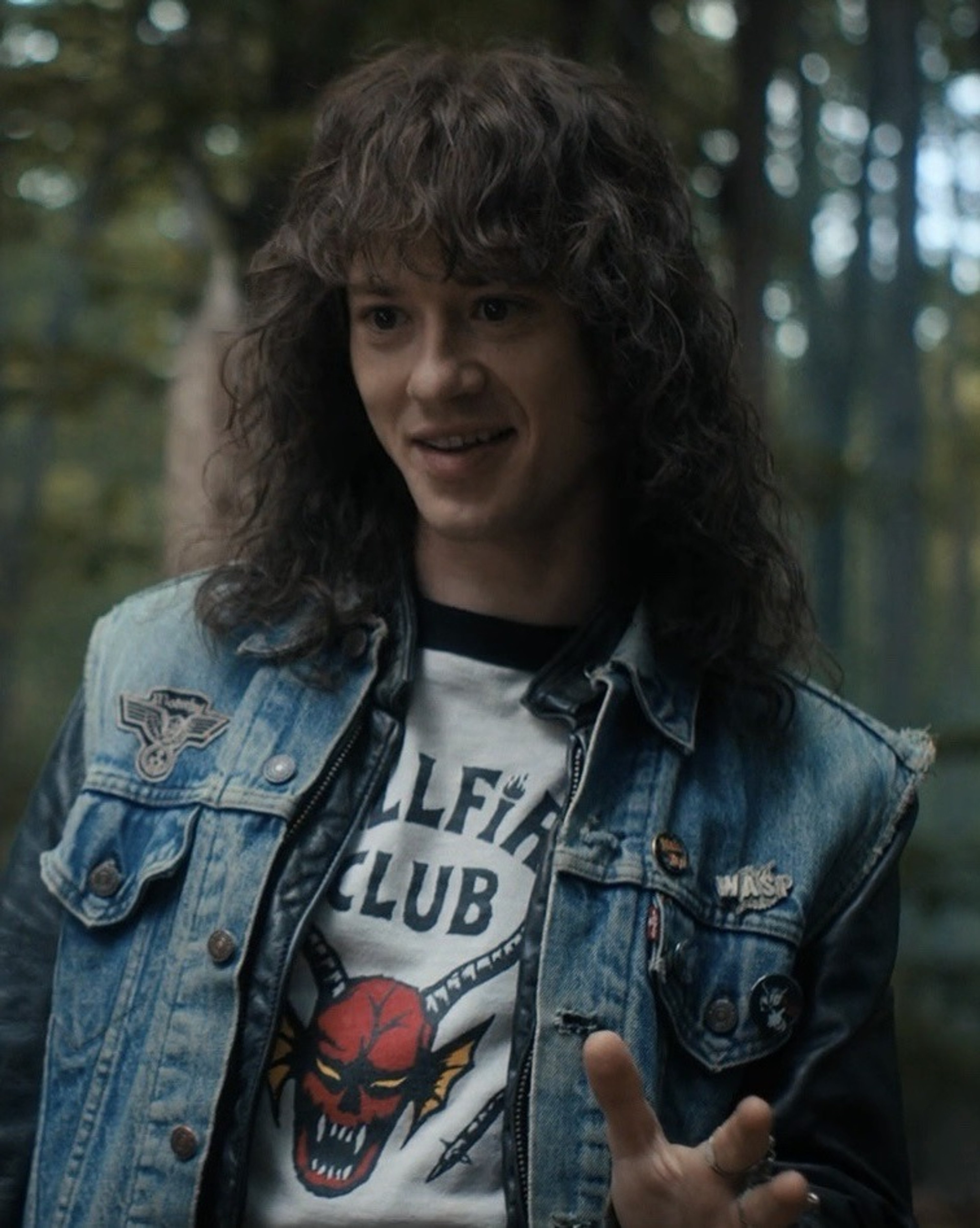 A young man with long, curly brown hair wearing a denim jacket and t-shirt in a forested setting.