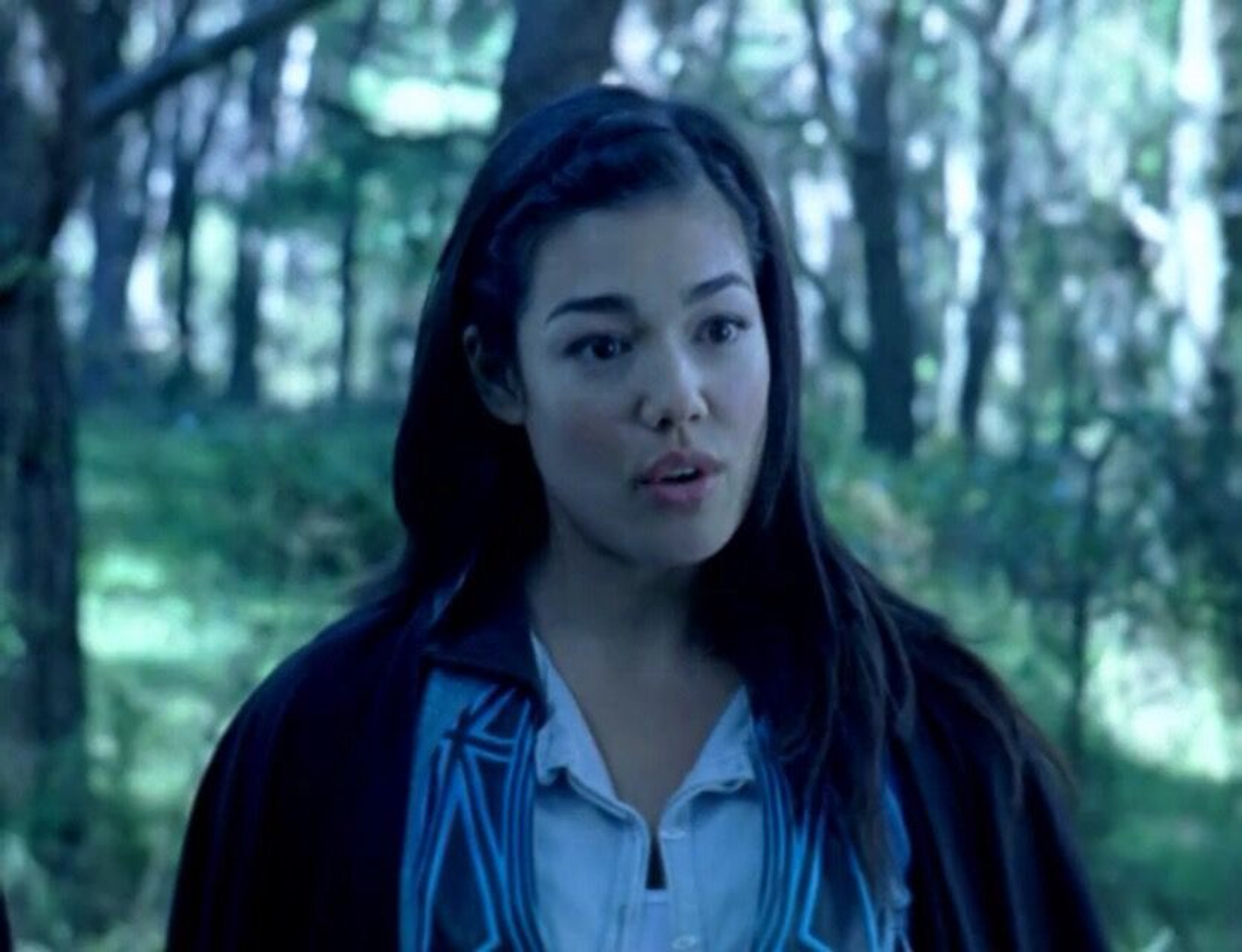 A young woman with long dark hair wearing a blue jacket in a wooded area