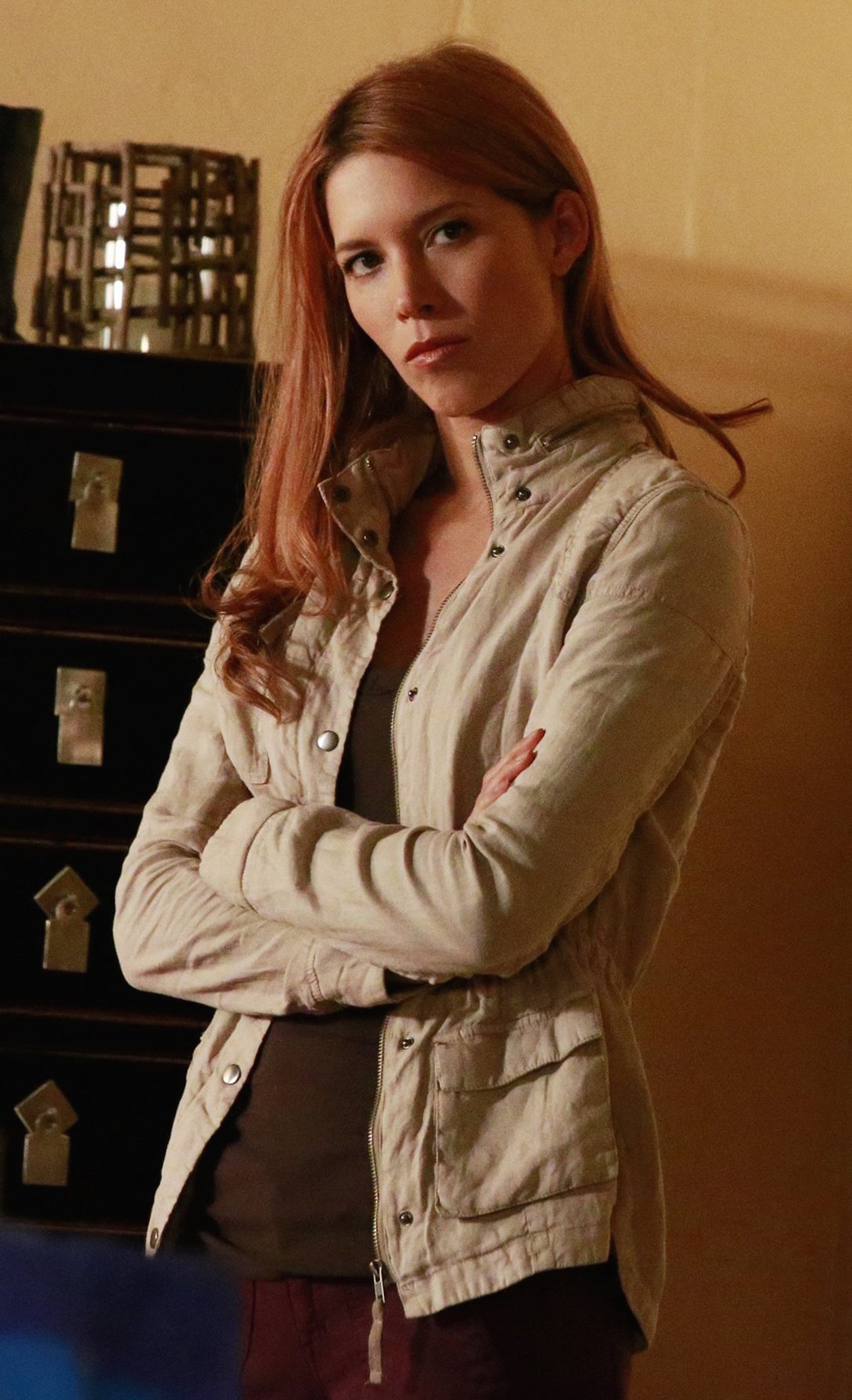 A young woman with red hair wearing a beige jacket, looking serious with her arms crossed.