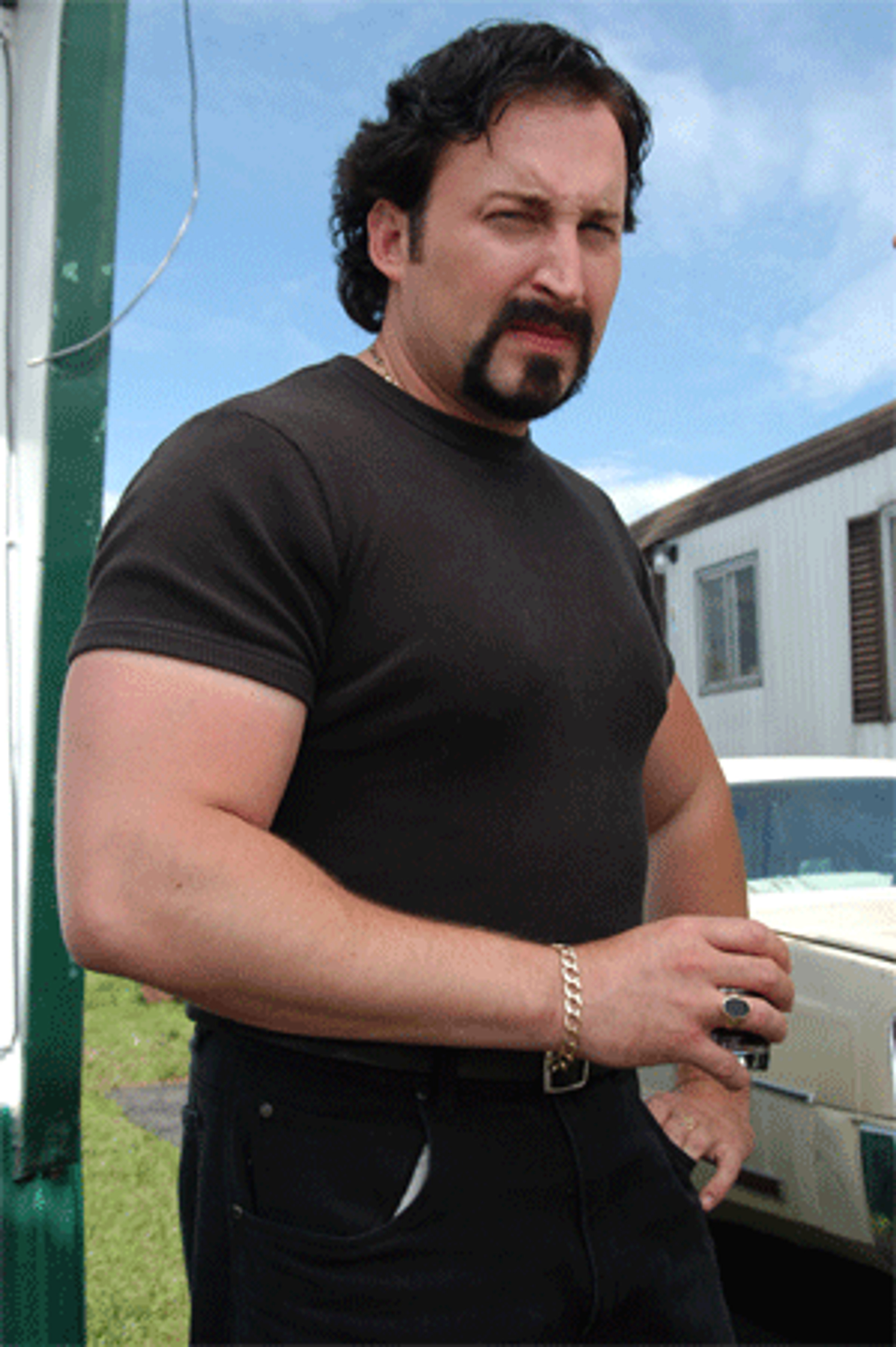 A middle-aged man with a muscular build and a beard, wearing a black t-shirt and standing outdoors.
