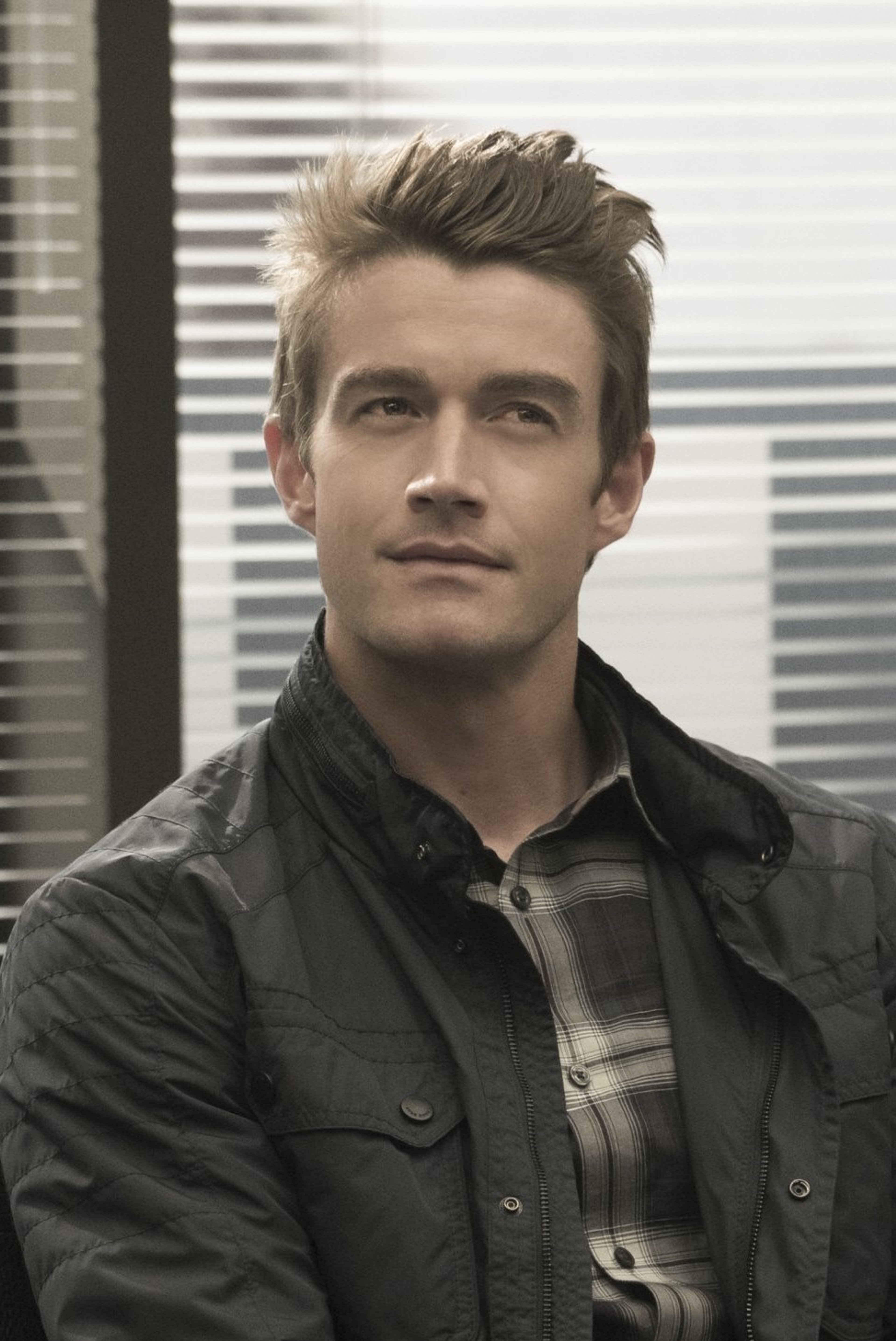 A man with short blonde hair and a serious expression, wearing a dark jacket and plaid shirt.