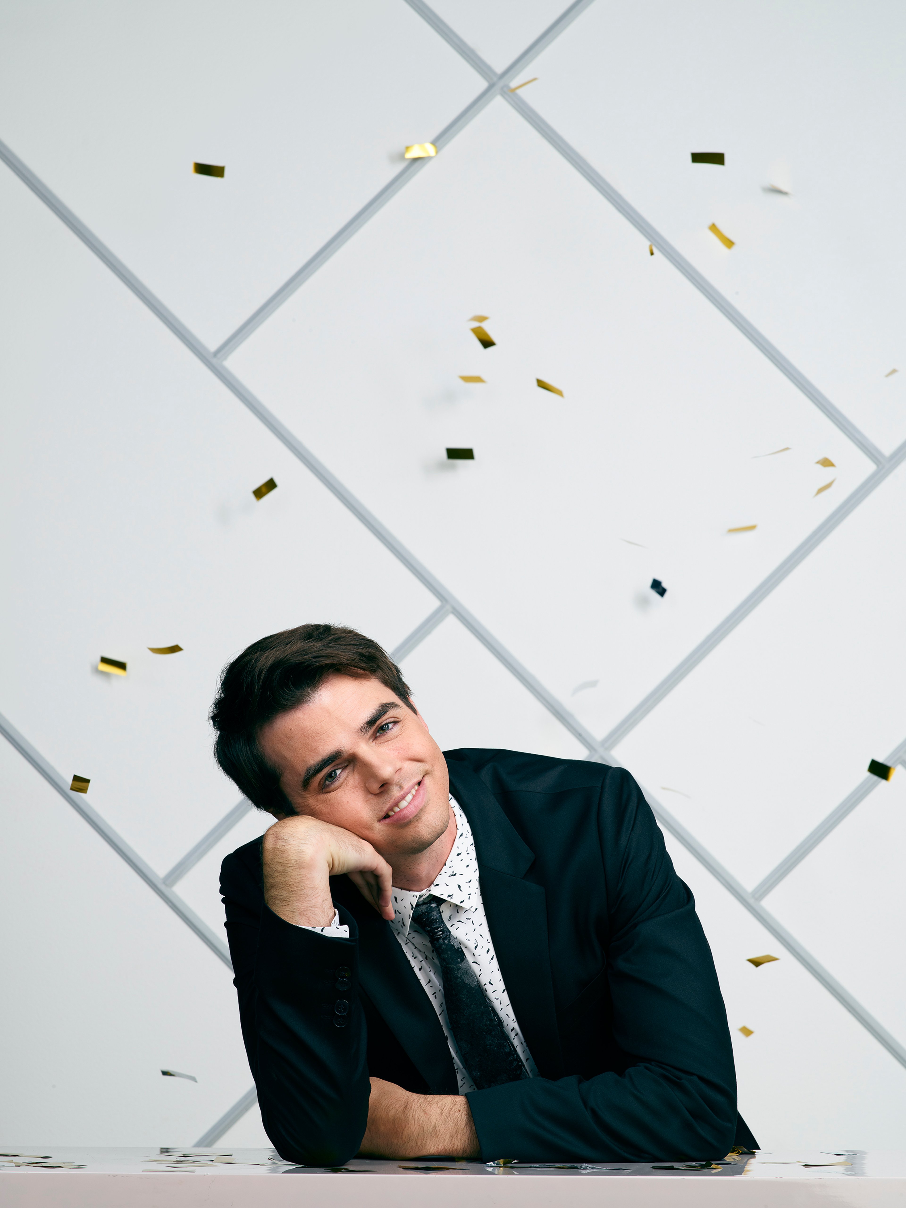A young man in a black suit and tie with a serious expression