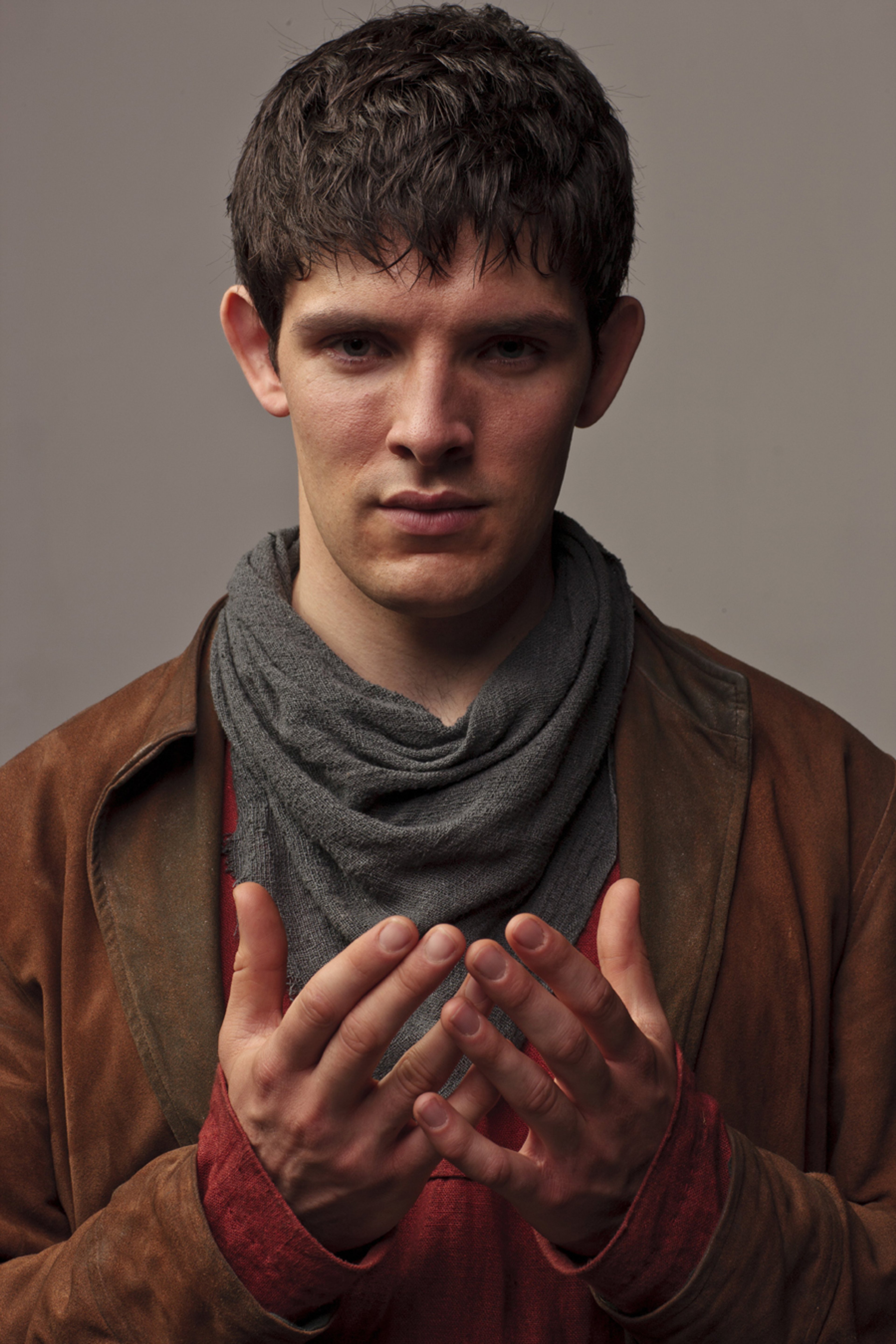 A young man with dark hair wearing a brown jacket and gray scarf