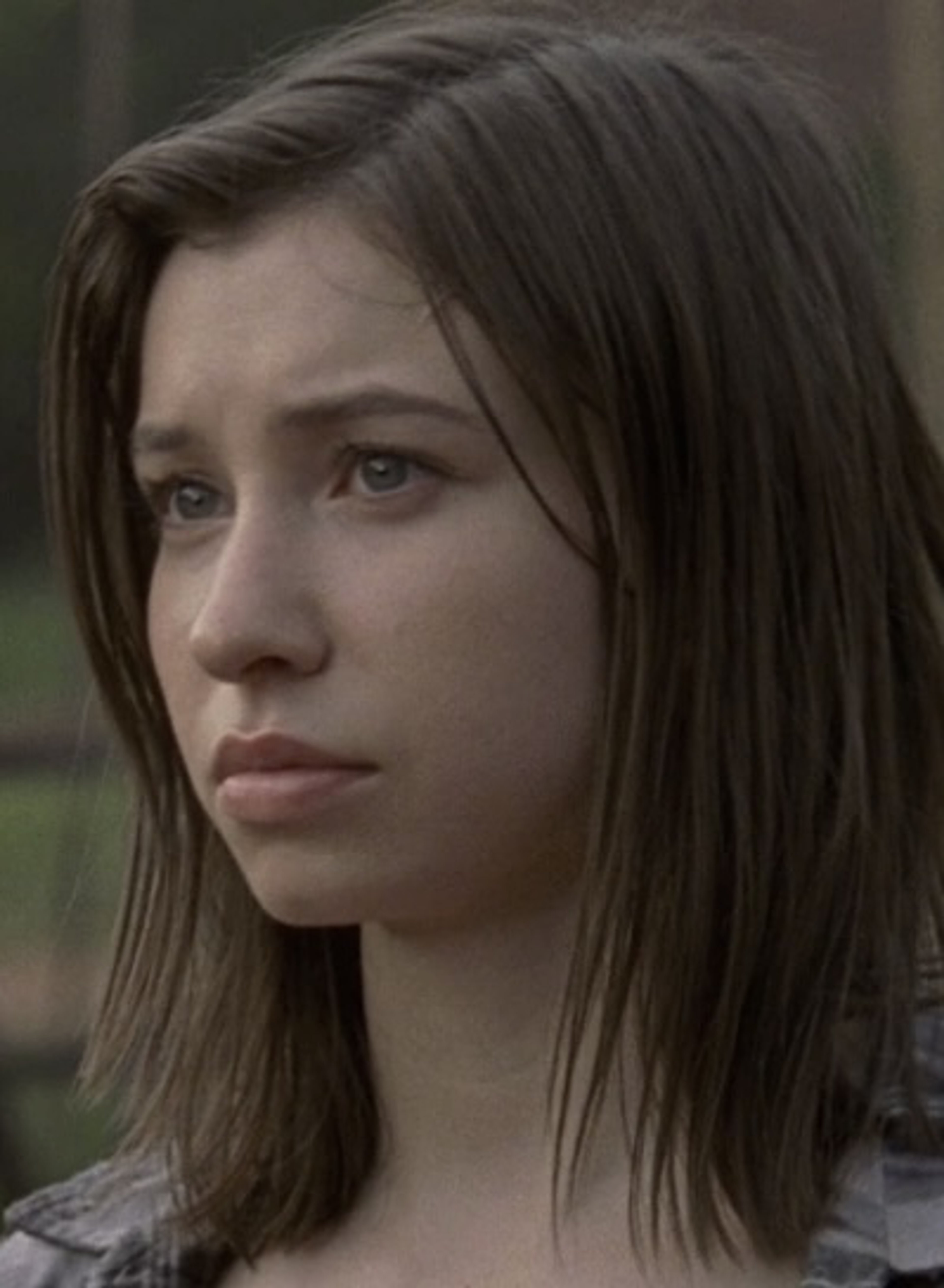 A young woman with dark hair in a serious, cynical expression against a bleak, gray background