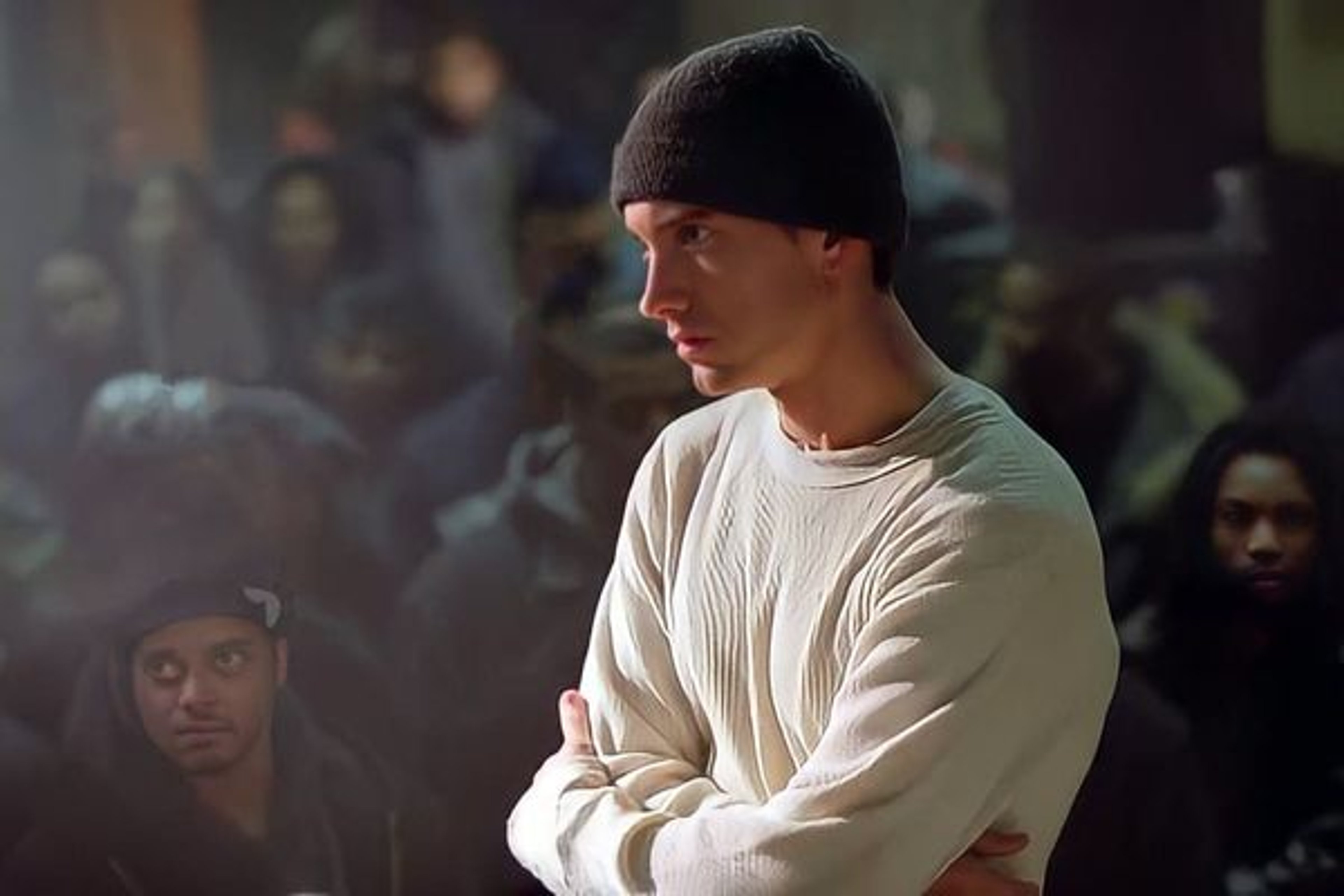 A young white man with short dark hair wearing a white hooded sweatshirt in a dimly lit setting.