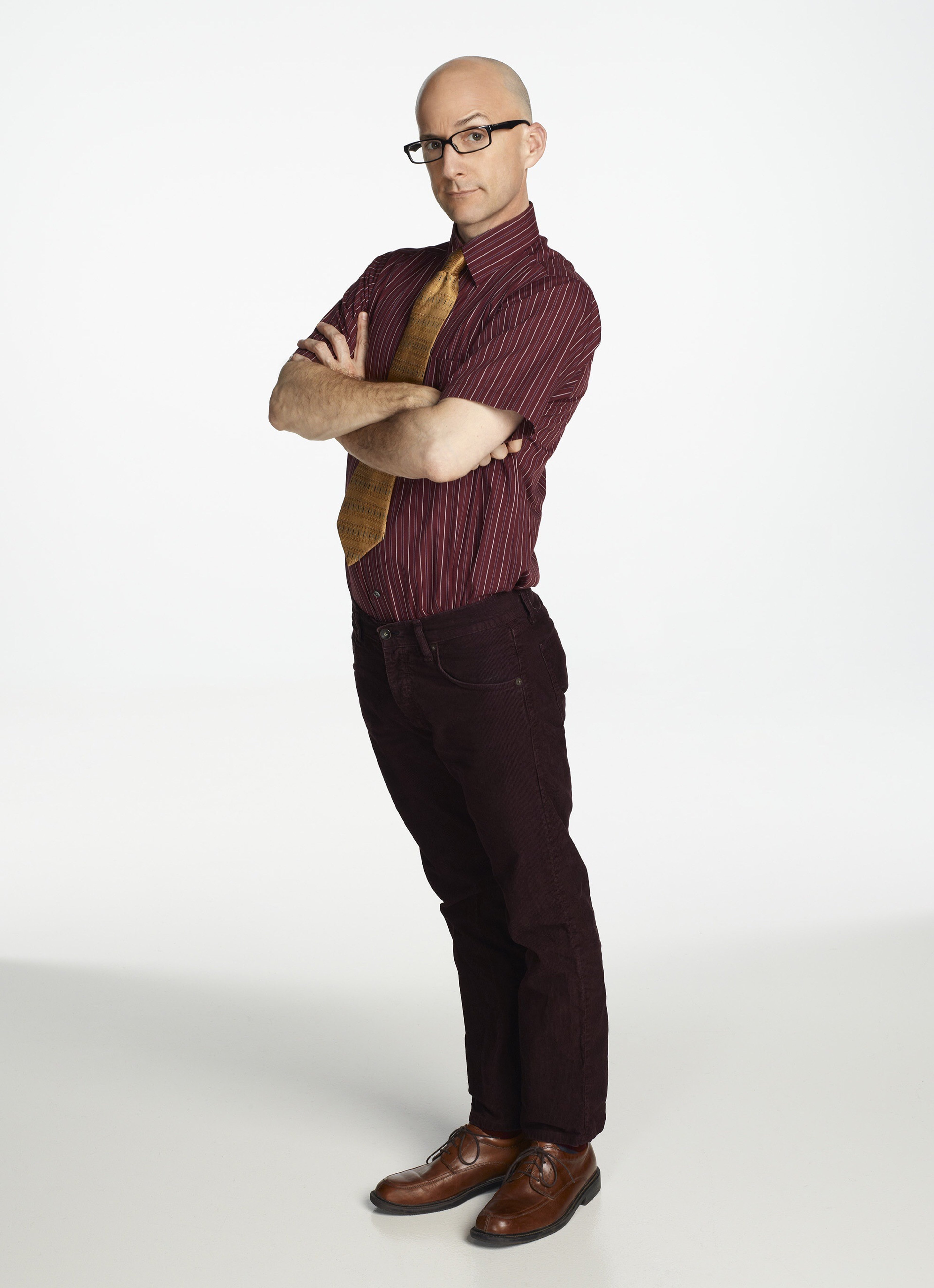 A middle-aged bald man in a burgundy shirt and tie with a serious expression, standing with his arms crossed.