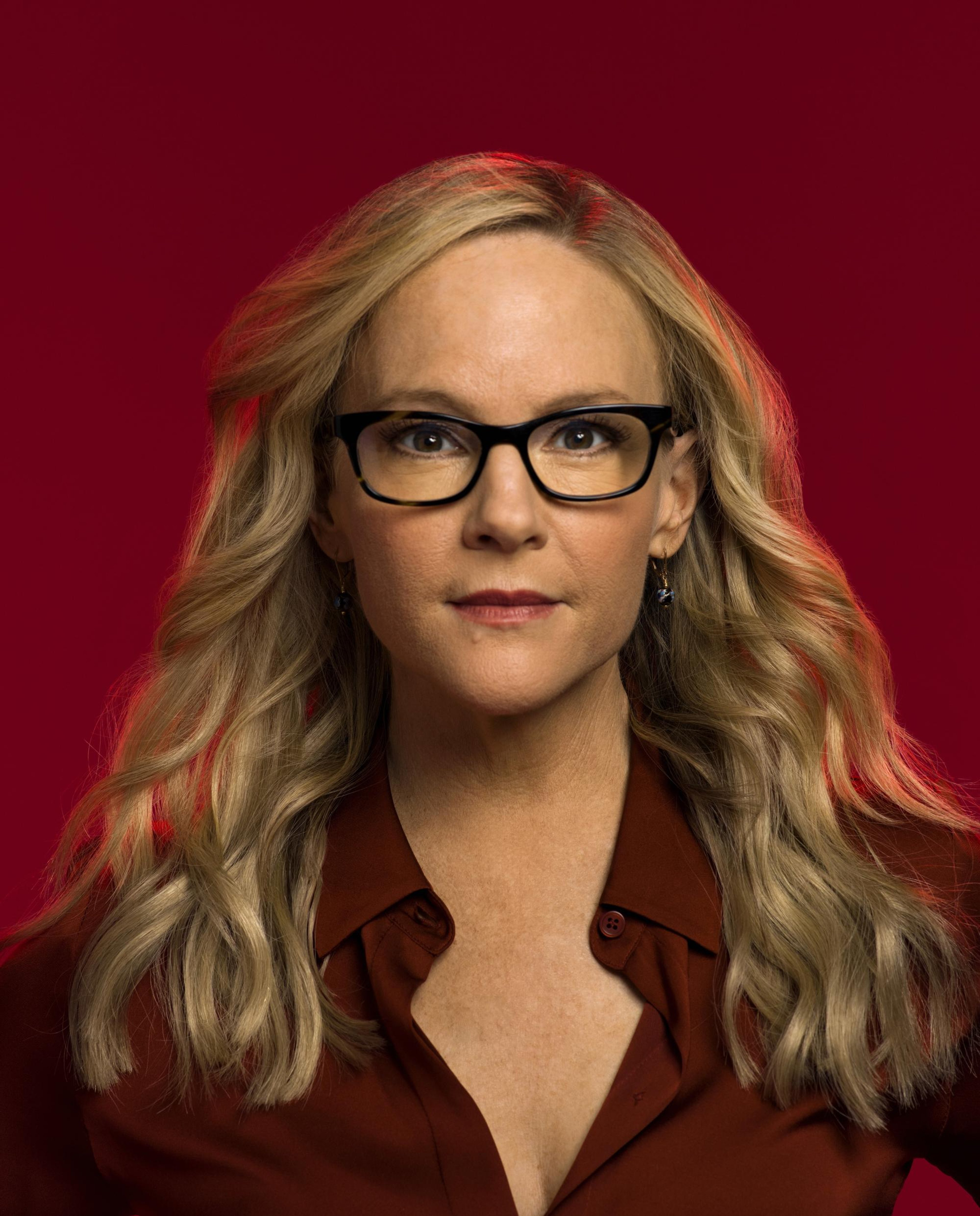 A woman with blonde hair and glasses wearing a brown jacket against a red background