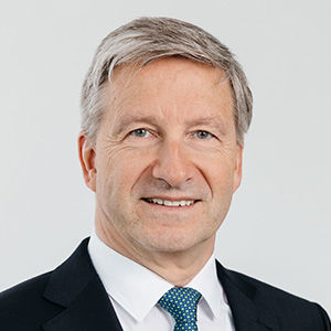 Headshot of Axel Stepken, a midle-aged man in a suit on a simple light gray background.