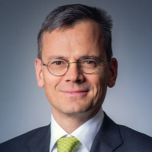 Headshot of Dominik Asam, a midle-aged man in a suit on a simple blue-gray background.