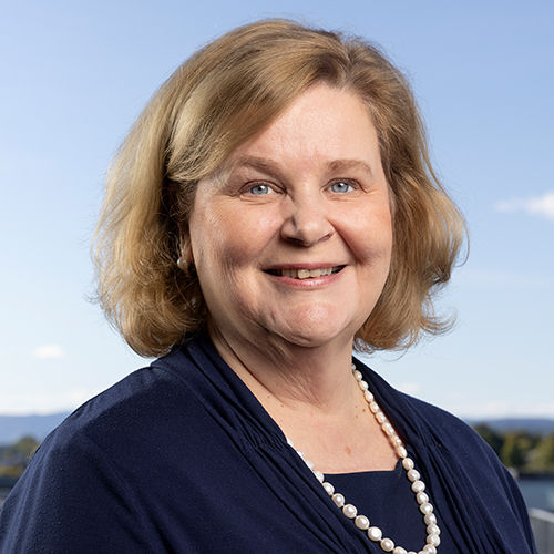 Headshot of Karen Spens, a midle-aged woman in a custume. In the background you can see the sky and a landscape.