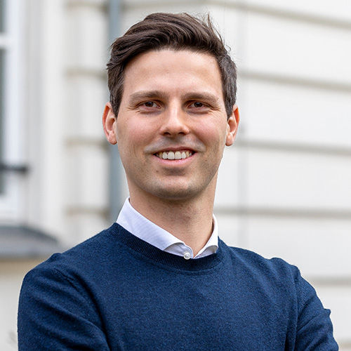 Prof. Jochen Hartmann, a young man, stands in front of a white building and is smiling.