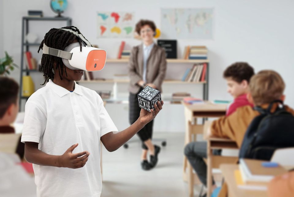 Student stood in classroom among fellow students, wearing a VR headset and holding an AR cube