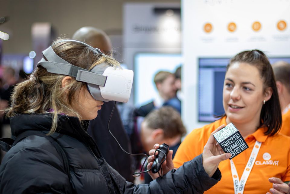 A person wearing a VR headset holding an AR cube, as a ClassVR colleague passes them a controller