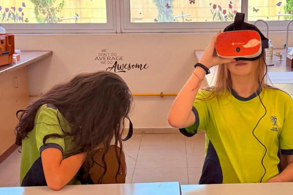 Two students sat at a desk, one with their head down, the other looking up wearing a VR headset