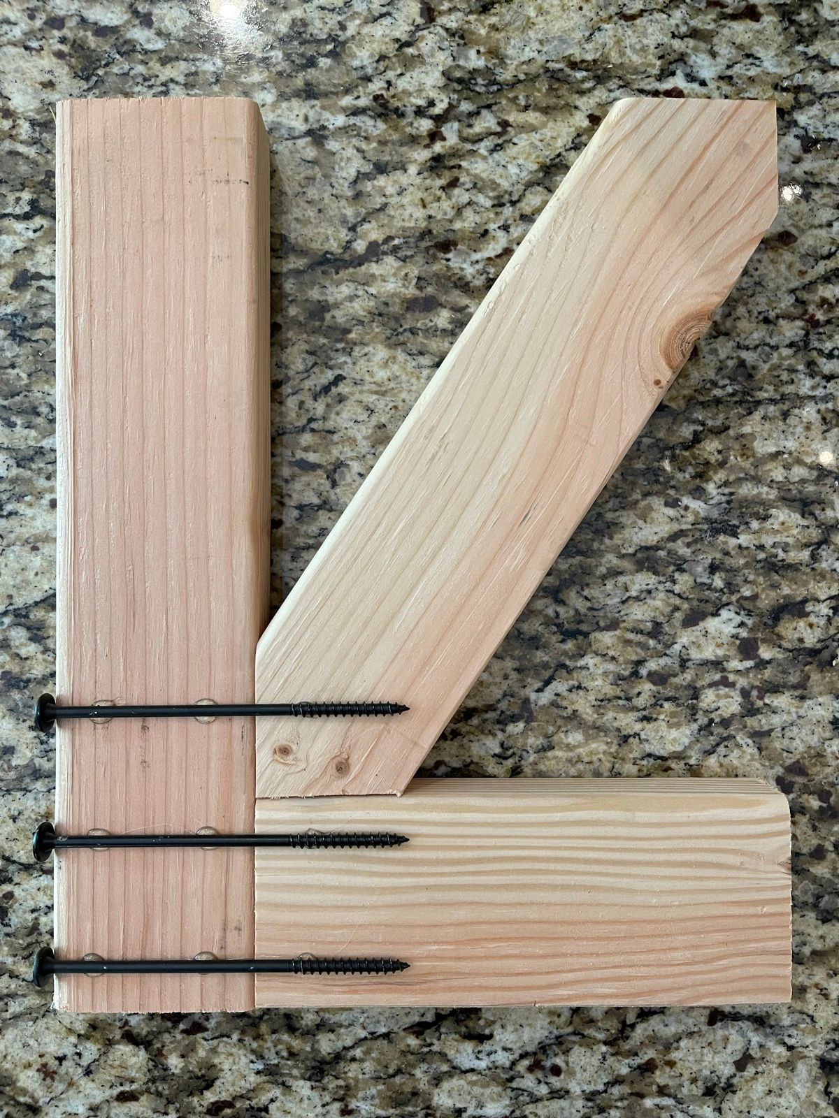 Corner joint of cedar gate frame showing horizontal rail, vertical post, and diagonal brace secured with long black structural screws from underneath