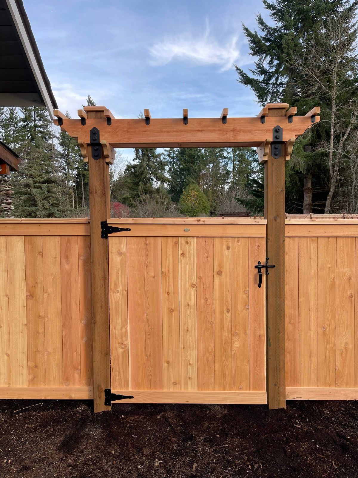 Cedar gate with arbor and black strap hinges, horizontal cedar planks, evergreen trees and blue sky