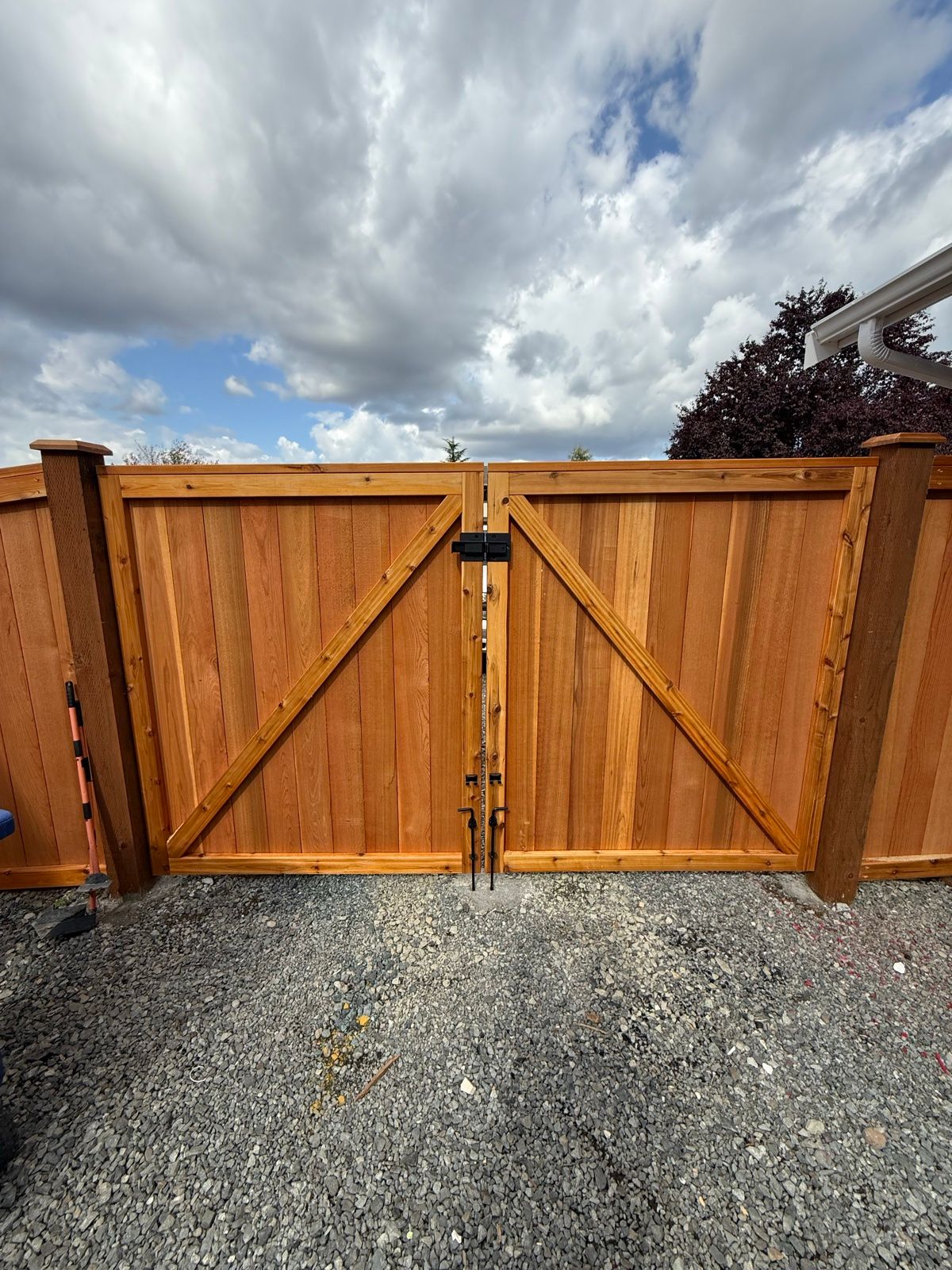 Double driveway gate with diagonal bracing, black latch hardware, gravel driveway and ornamental tree