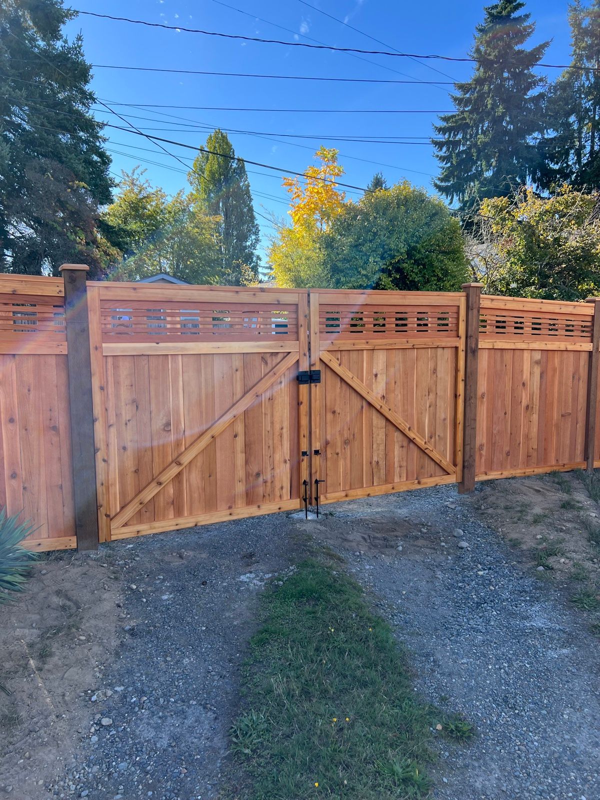 Double driveway gate with Z-bracing and lattice-top fence above, gravel driveway, trees and power lines in background