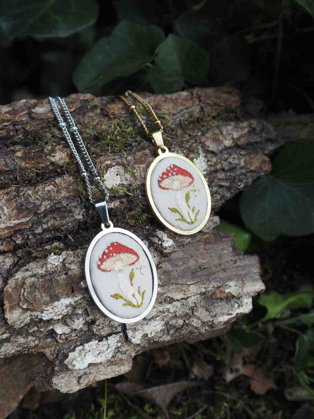 L'image présente deux colliers pendentifs ovales, chacun orné d'une illustration de champignon amanite tue-mouches, posés sur un morceau de bois flotté ou d'écorce. Voici une description détaillée : *   **Composition générale :** Les deux colliers sont disposés côte à côte sur un support naturel, probablement un tronc d'arbre ou une grosse branche, dont l'écorce est visible et texturée. L'arrière-plan est flou, suggérant un environnement extérieur avec de la verdure (feuilles de lierre ou autres plantes grimpantes) et de la terre ou de la mousse.
*   **Les colliers :**
    *   **Collier de gauche :** Il a une chaîne et un cadre de pendentif de couleur argentée. Le pendentif est ovale et contient une illustration détaillée d'un champignon amanite tue-mouches. Le champignon a un chapeau rouge vif parsemé de points blancs, et un pied blanc avec des détails verts (herbe ou mousse) à sa base. Le fond de l'illustration est clair, probablement blanc cassé ou crème.
    *   **Collier de droite :** Il a une chaîne et un cadre de pendentif de couleur dorée. Le pendentif est également ovale et présente une illustration similaire du même type de champignon, avec un chapeau rouge à points blancs et un pied blanc avec des éléments verts à la base. Le fond de l'illustration est également clair.
*   **Le support :** Le bois sur lequel reposent les colliers est rugueux et texturé, avec des crevasses et des motifs naturels de l'écorce. Sa couleur est principalement marron clair à grisâtre.
*   **Lumière et ambiance :** La lumière semble naturelle, peut-être un peu tamisée, ce qui met en valeur les couleurs des pendentifs et crée une atmosphère douce et naturelle. L'arrière-plan flou aide à focaliser l'attention sur les bijoux. En résumé, l'image met en scène deux bijoux artisanaux ou fantaisie, inspirés de la nature et du monde des champignons, présentés de manière esthétique dans un cadre naturel.