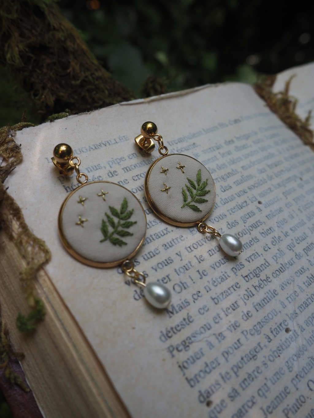 L'image montre une paire de boucles d'oreilles élégantes posées sur un livre ouvert. Les boucles d'oreilles sont composées de médaillons ronds blancs décorés de motifs de fougères vertes et de petites étoiles dorées. Au bas de chaque médaillon pend une petite perle nacrée. Les boucles d'oreilles semblent être montées avec des clips ou des tiges dorées. L'arrière-plan montre les pages d'un livre ancien avec du texte en français, donnant à l'image une ambiance vintage et romantique. La composition est artistique, avec un éclairage doux et naturel qui met en valeur les détails délicats des bijoux. Ces boucles d'oreilles ont un style botanique et délicat qui évoque la nature et le printemps, avec leur design incorporant des éléments naturels comme les fougères.