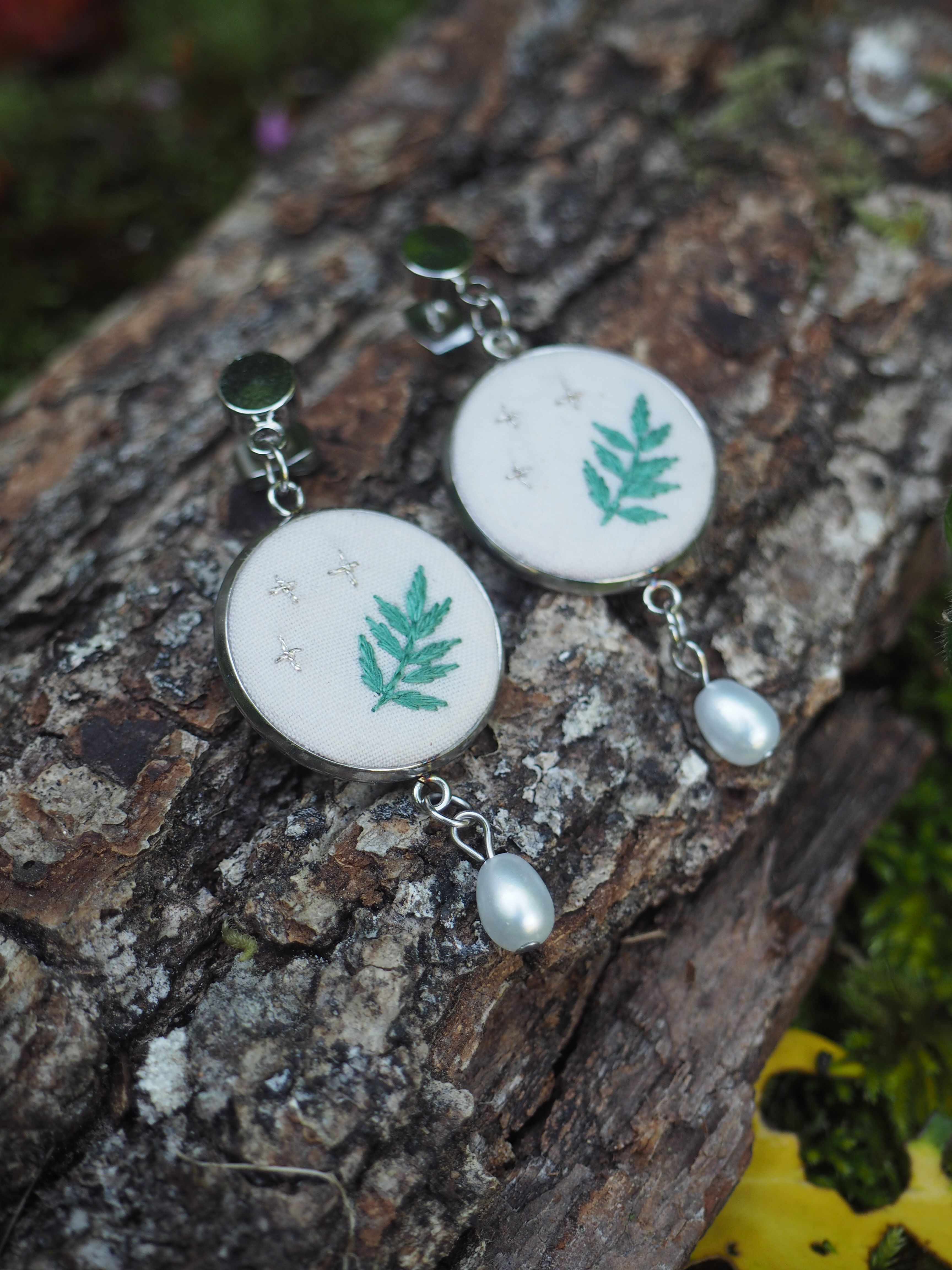 Cette image montre une paire de boucles d'oreilles artisanales posées sur un morceau d'écorce d'arbre. Les boucles d'oreilles sont composées de médaillons blancs circulaires décorés d'une feuille de fougère turquoise. Au bas de chaque médaillon pend une petite perle nacrée. Les boucles d'oreilles semblent être faites en argent ou en métal argenté. L'arrière-plan naturel avec l'écorce texturée et un peu de mousse verte crée une belle mise en scène qui met en valeur ces bijoux délicats inspirés de la nature.