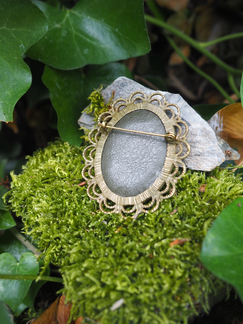 L'image montre des broches ou des bijoux vintage avec des motifs floraux décoratifs en métal doré, disposés sur un lit de mousse verte. Les broches semblent avoir des centres argentés ou gris mat, et leur design rappelle des fleurs stylisées avec des bordures ornementales. Le fond présente également quelques feuilles vertes, créant un cadre naturel et organique pour ces pièces de bijouterie délicates. L'ambiance de la photo est très romantique et évoque un style vintage ou antique.
