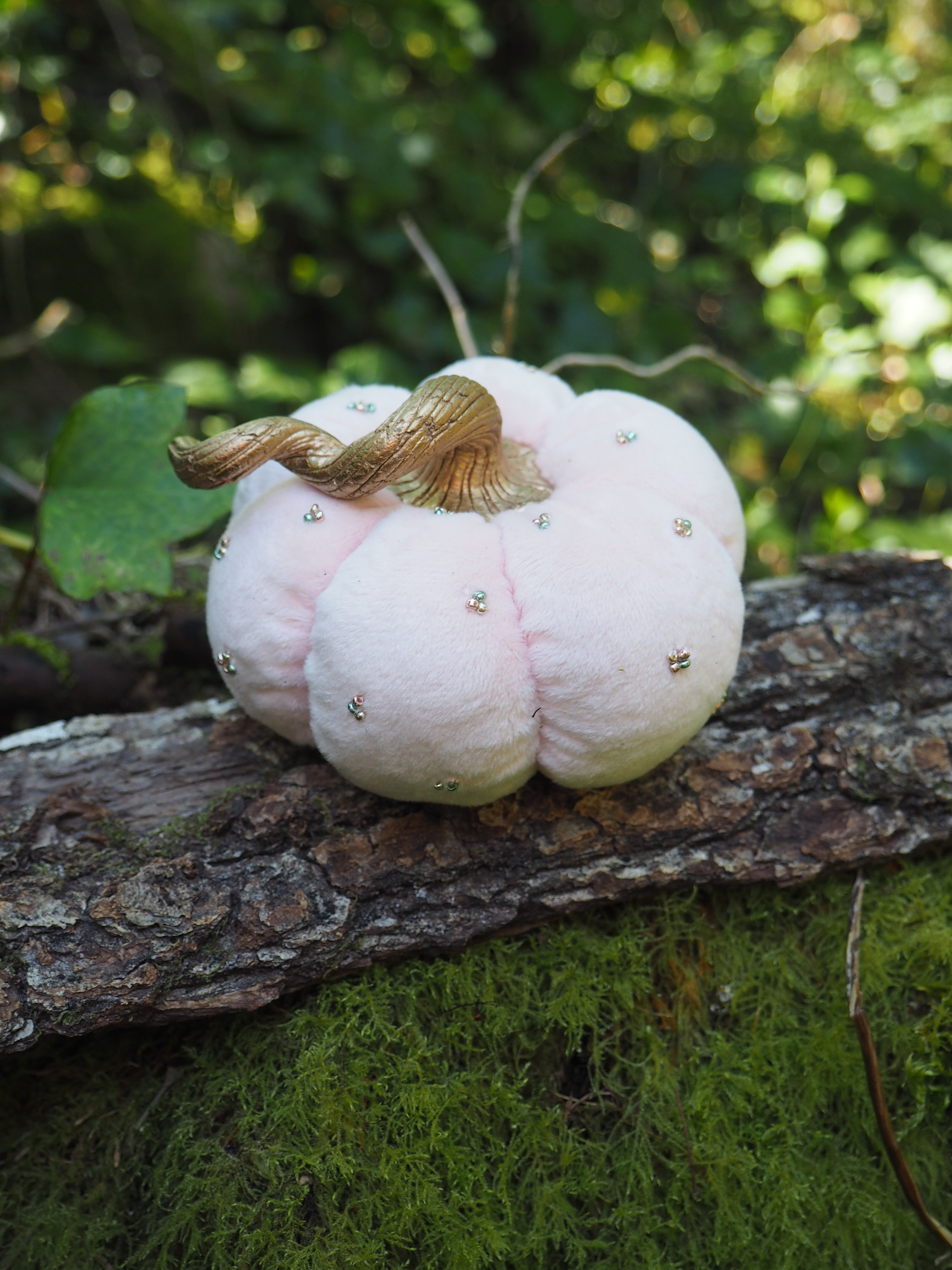 Cette image montre une création artistique qui ressemble à une petite citrouille ou courge décorative faite en tissu rose pâle ou blanc. Elle est placée sur un morceau de bois couvert de mousse verte. La citrouille est ornée de petits détails dorés ou métalliques et possède ce qui semble être une tige ou une feuille naturelle séchée sur le dessus. L'arrière-plan est flou mais on peut distinguer des feuilles vertes, donnant une ambiance forestière ou de jardin à la scène. C'est une composition très délicate et poétique qui mélange des éléments naturels et artificiels.