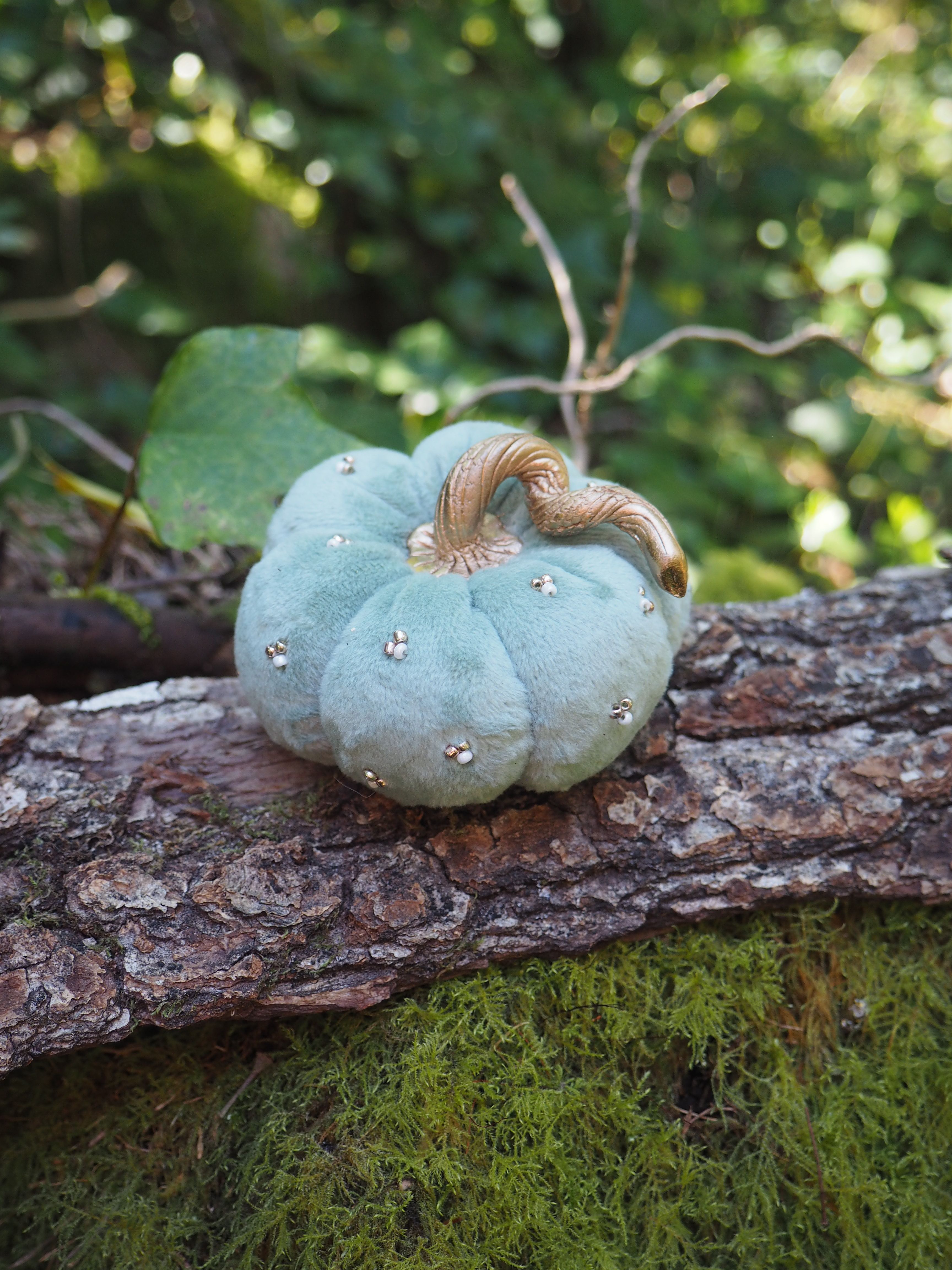 Cette image montre une citrouille décorative dans un style fantaisiste. Elle est de couleur bleu pâle ou vert d'eau, avec une tige naturelle de couleur cuivrée. La citrouille est ornée de petits strass ou cristaux décoratifs qui scintillent. Elle est placée sur un tronc d'arbre ou une branche couverte de mousse verte, créant une ambiance féerique et enchantée. L'arrière-plan est flou mais on peut distinguer un environnement naturel verdoyant. Cette décoration s'inscrit dans un style "cottage core" ou féérique, rappelant un peu l'univers de Cendrillon mais avec une touche moderne et élégante.