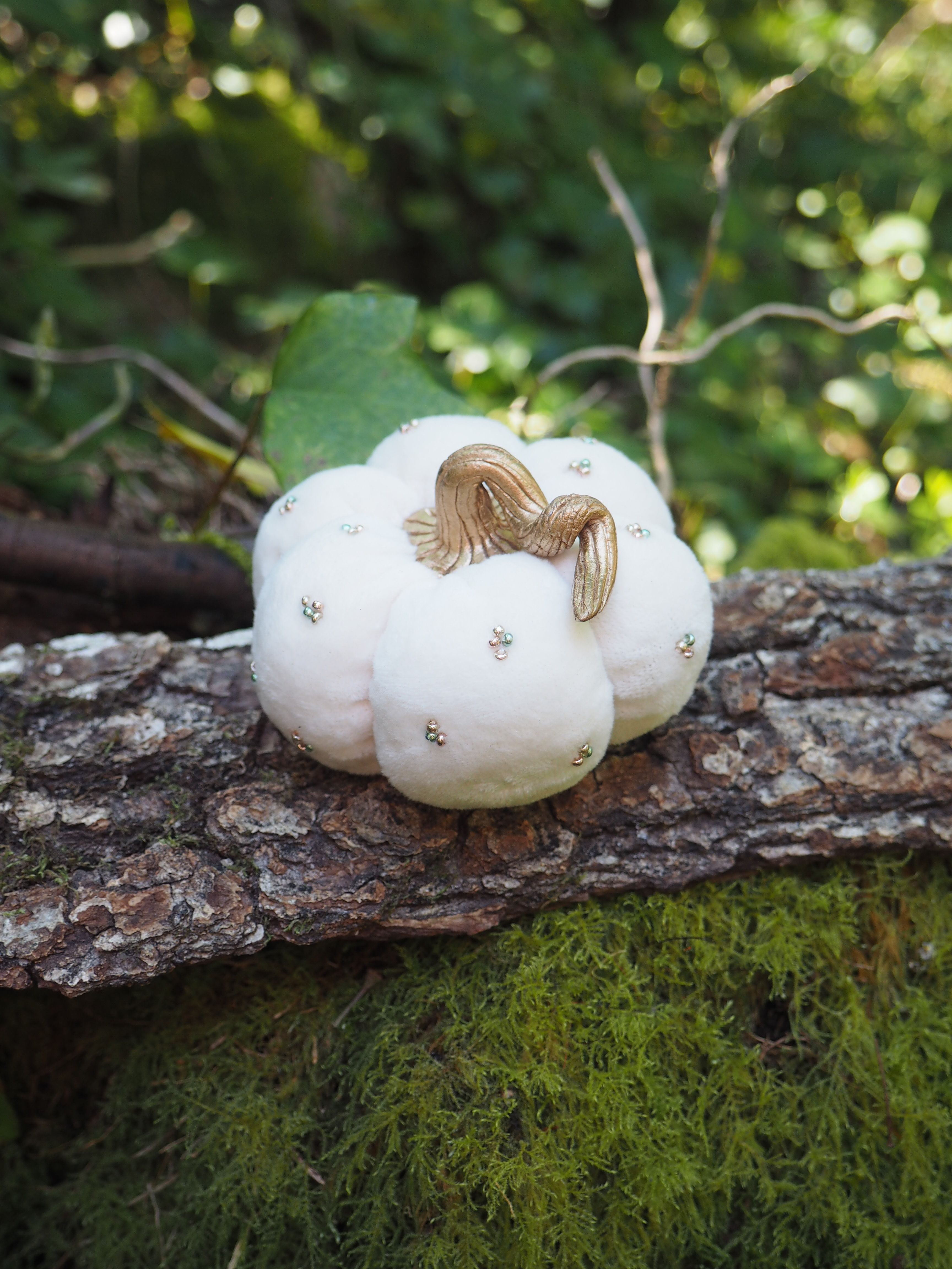 Cette image montre une citrouille décorative blanche, probablement faite de tissu ou de matériau similaire, posée sur un tronc d'arbre ou une branche couverte de mousse verte. La citrouille a une tige dorée ou bronze et semble être une décoration automnale ou d'Halloween. L'arrière-plan est verdoyant avec un effet de bokeh (flou artistique) créé par la lumière filtrant à travers les feuilles. C'est une composition très naturelle et paisible qui mélange des éléments décoratifs avec la nature.