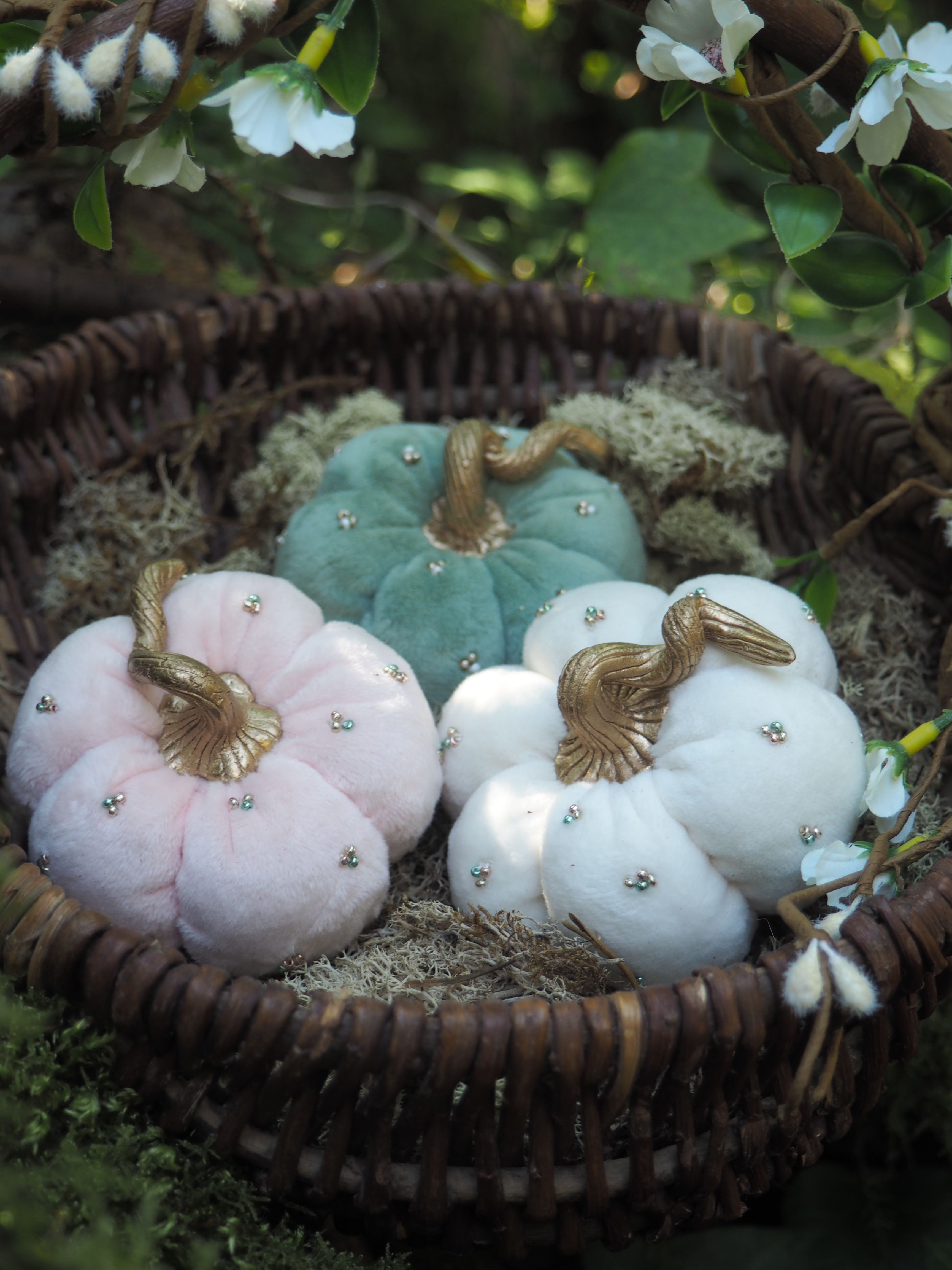 Cette image montre trois jolies citrouilles décoratives en tissu ou en feutre, disposées dans un panier en osier foncé. Les citrouilles sont de style shabby chic ou vintage, dans des tons pastel : une rose pâle, une bleu turquoise et une blanche. Chacune est ornée d'une tige métallique dorée qui ressemble à un oiseau ou à un motif floral. Les citrouilles sont également décorées de petites perles ou strass qui ajoutent une touche scintillante. Le panier est placé dans un cadre naturel avec des feuilles vertes et des fleurs blanches en arrière-plan, créant une ambiance douce et romantique. C'est une décoration parfaite pour l'automne qui combine élégance et douceur.