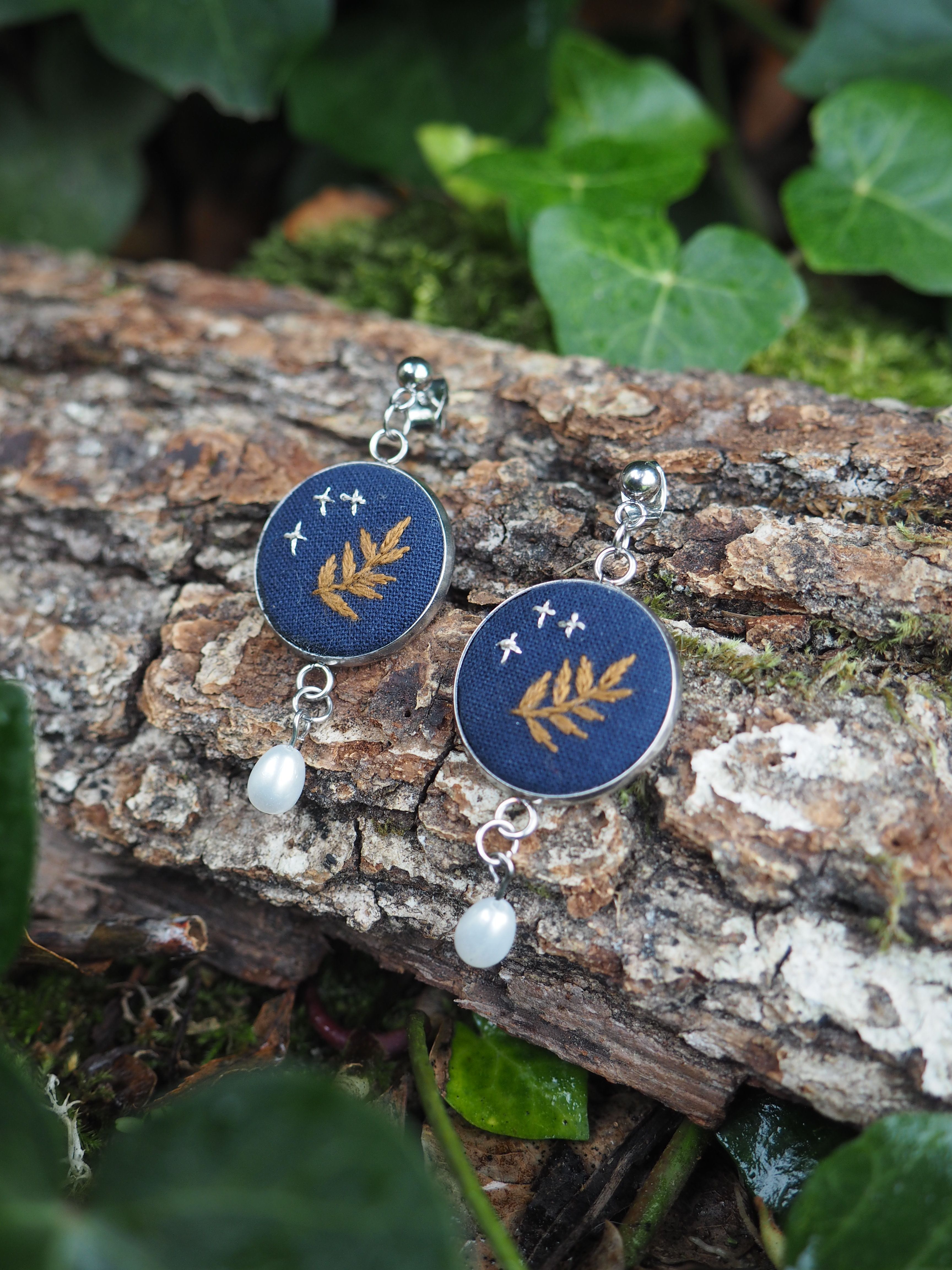 Cette image présente une paire de boucles d'oreilles artisanales disposées sur un morceau de bois naturel dans un environnement végétal. **Description des bijoux :**
- Boucles d'oreilles avec des pendentifs circulaires de couleur bleu foncé/marine
- Chaque pendentif est orné d'un motif doré représentant une feuille ou une branche délicate
- De petites étoiles dorées parsèment le fond bleu, créant un effet céleste
- Une perle blanche nacrée pend élégamment sous chaque pendentif principal
- Les attaches et chaînettes sont en métal argenté **Mise en scène :**
- Les bijoux reposent sur une branche ou un tronc d'arbre à l'écorce rugueuse et naturelle
- L'arrière-plan est composé de feuillage vert luxuriant, légèrement flou
- L'éclairage naturel met en valeur les détails dorés et la brillance des perles
- Cette présentation évoque un style bohème et naturel, soulignant le caractère artisanal des pièces L'ensemble dégage une esthétique poétique mêlant thèmes botaniques et cosmiques, parfaite pour des bijoux de créateur inspirés de la nature.