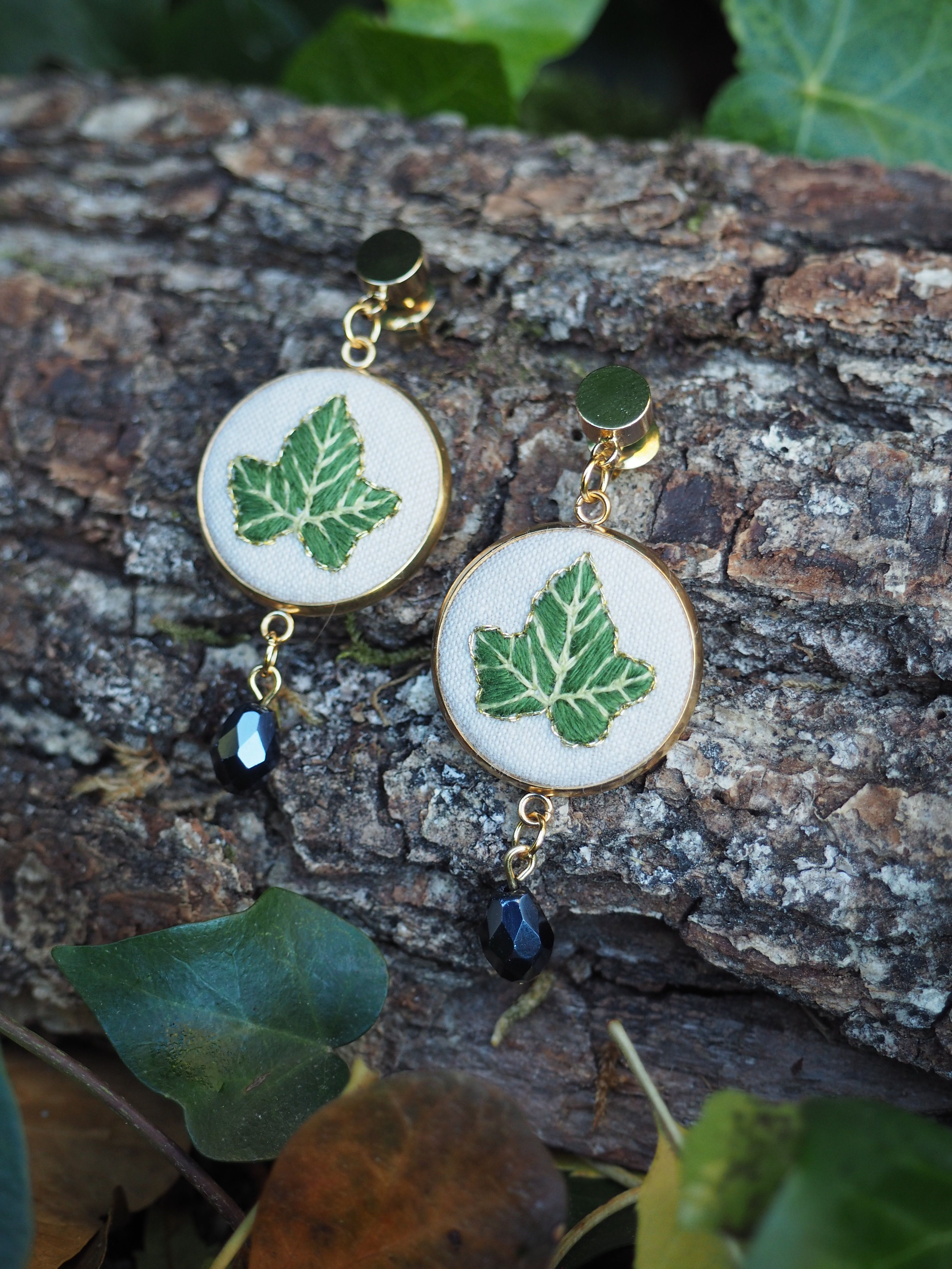 Cette image présente une paire de boucles d'oreilles artisanales disposées sur un tronc d'arbre dans un environnement naturel.  **Caractéristiques des bijoux :**
- Pendants circulaires en résine ou matériau similaire avec un fond blanc/crème
- Motifs de feuilles vertes délicatement incrustés ou peints au centre de chaque pendentif
- Montures dorées avec des crochets d'oreilles assortis
- Petites perles pendantes de couleur bleu foncé/noir qui complètent l'ensemble **Mise en scène :**
- Les boucles d'oreilles reposent sur l'écorce rugueuse et texturée d'un tronc d'arbre
- L'environnement naturel est souligné par la présence de vraies feuilles vertes disposées autour des bijoux
- L'éclairage naturel met en valeur les détails et crée une atmosphère organique et harmonieuse Cette composition photographique met parfaitement en valeur le thème botanique des bijoux en les intégrant dans leur environnement d'inspiration naturelle.