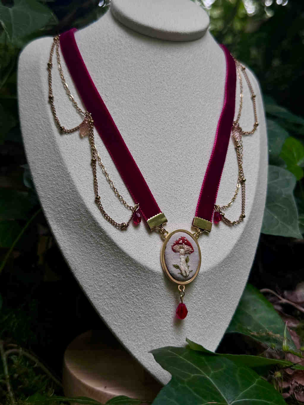 Cette image présente un collier de style choker, exposé sur un buste de présentation blanc, dans un environnement qui évoque une forêt ou un jardin luxuriant. Le collier est composé d'un ruban de velours de couleur bordeaux ou framboise, qui forme la partie principale du tour de cou. De chaque côté du ruban, des chaînes fines de couleur dorée sont drapées, créant un effet de cascade. Ces chaînes sont ornées de petites perles ou cristaux translucides de couleur rose pâle, ainsi que de perles rouges plus foncées près du centre. Au centre du collier, un pendentif circulaire est suspendu. Il est encadré d'un anneau doré et présente une broderie délicate sur un fond clair, représentant un champignon amanite muscaria avec son chapeau rouge à pois blancs et son pied blanc. Sous ce pendentif central, une perle rouge en forme de goutte est suspendue, ajoutant une touche finale. Le buste de présentation blanc contraste avec les couleurs riches du collier et l'environnement verdoyant. L'arrière-plan est flou, mais on distingue des feuilles vertes, probablement du lierre ou d'autres plantes de sous-bois, ce qui renforce l'esthétique naturelle et féerique de la scène. La lumière semble douce et naturelle, mettant en valeur les détails et les textures du collier.