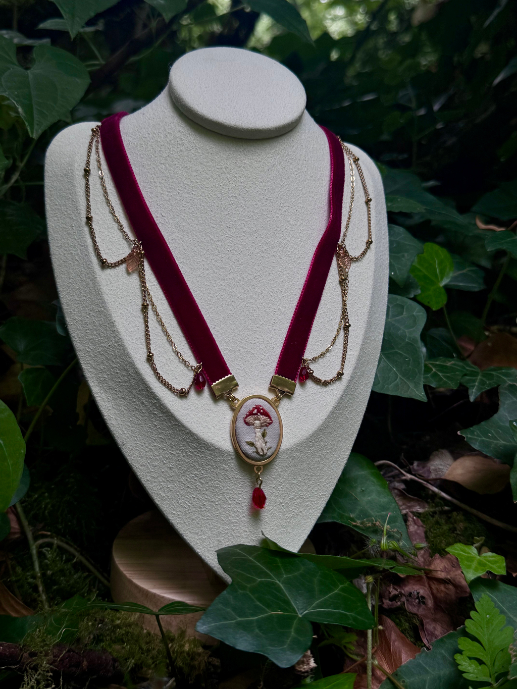 L'image présente un collier de style choker, exposé sur un présentoir de bijoux de couleur crème, sur un fond de verdure luxuriante. Le collier est composé d'un ruban de velours bordeaux foncé, qui forme la base du choker. De chaque côté du ruban, des chaînes fines de couleur or sont attachées, créant un effet de drapé élégant. Ces chaînes sont ornées de petites perles ou de breloques discrètes. Au centre du collier, un pendentif circulaire attire l'attention. Il est encadré d'un anneau doré et présente une broderie délicate d'un champignon rouge et blanc, rappelant un amanite tue-mouches, sur un fond clair. Sous ce pendentif central, une petite breloque en forme de goutte de couleur rouge foncé pend, ajoutant une touche de couleur et de mouvement. Le présentoir sur lequel repose le collier est simple et met en valeur le bijou. Il est posé sur une surface en bois clair, qui est elle-même entourée de feuilles vertes et de mousse, suggérant un environnement naturel, peut-être une forêt ou un jardin. La lumière naturelle éclaire la scène, créant des ombres douces et accentuant les textures. L'ensemble de l'image dégage une atmosphère féerique et naturelle, le collier semblant s'intégrer parfaitement dans ce décor végétal.