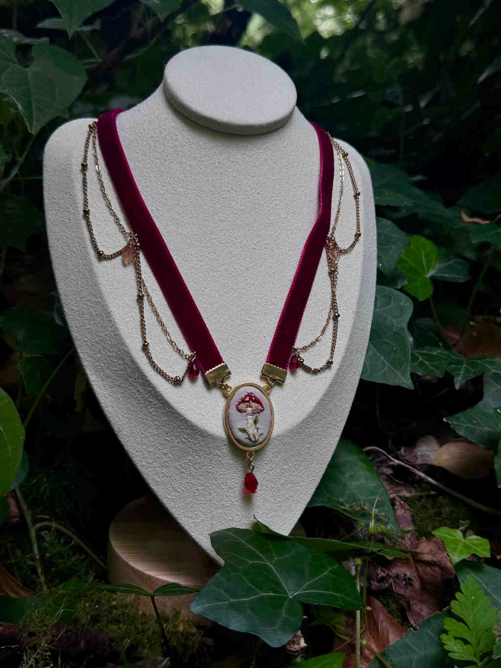 L'image présente un collier de style choker, exposé sur un présentoir de bijoux de couleur crème, sur un fond de verdure luxuriante. Le collier est composé d'un ruban de velours bordeaux foncé, qui forme la base du choker. De chaque côté du ruban, des chaînes fines de couleur or sont attachées, créant un effet de drapé élégant. Ces chaînes sont ornées de petites perles ou de breloques discrètes. Au centre du collier, un pendentif circulaire attire l'attention. Il est encadré d'un anneau doré et présente une broderie délicate d'un champignon rouge et blanc, rappelant un amanite tue-mouches, sur un fond clair. Sous ce pendentif central, une petite breloque en forme de goutte de couleur rouge foncé pend, ajoutant une touche de couleur et de mouvement. Le présentoir sur lequel repose le collier est simple et met en valeur le bijou. Il est posé sur une surface en bois clair, qui est elle-même entourée de feuilles vertes et de mousse, suggérant un environnement naturel, peut-être une forêt ou un jardin. La lumière naturelle éclaire la scène, créant des ombres douces et accentuant les textures. L'ensemble de l'image dégage une atmosphère féerique et naturelle, le collier semblant s'intégrer parfaitement dans ce décor végétal.