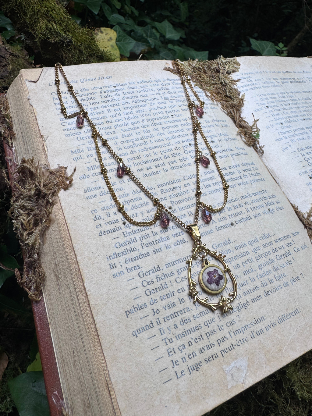 This image features a close-up, high-angle shot of a vintage-style necklace resting on an open, aged book. The necklace is made of a gold-toned metal chain, adorned with small, faceted purple beads interspersed along its length. The central pendant is a teardrop-shaped frame, also in gold-toned metal, with intricate scrollwork details. Inside the frame, there's a small, circular element with a purple flower design, possibly made of enamel or a similar material. The necklace is draped across the pages of the book, with parts of the chain extending off the edges. The book itself appears old, with yellowed pages and a slightly worn texture. The text on the pages is in French, printed in black ink. The book is open, revealing two full pages of text. The background and surrounding elements suggest an outdoor, natural setting. There are hints of green moss and dark foliage around the book, creating a rustic and somewhat mystical atmosphere. The lighting is soft, highlighting the details of the necklace and the texture of the book pages.
