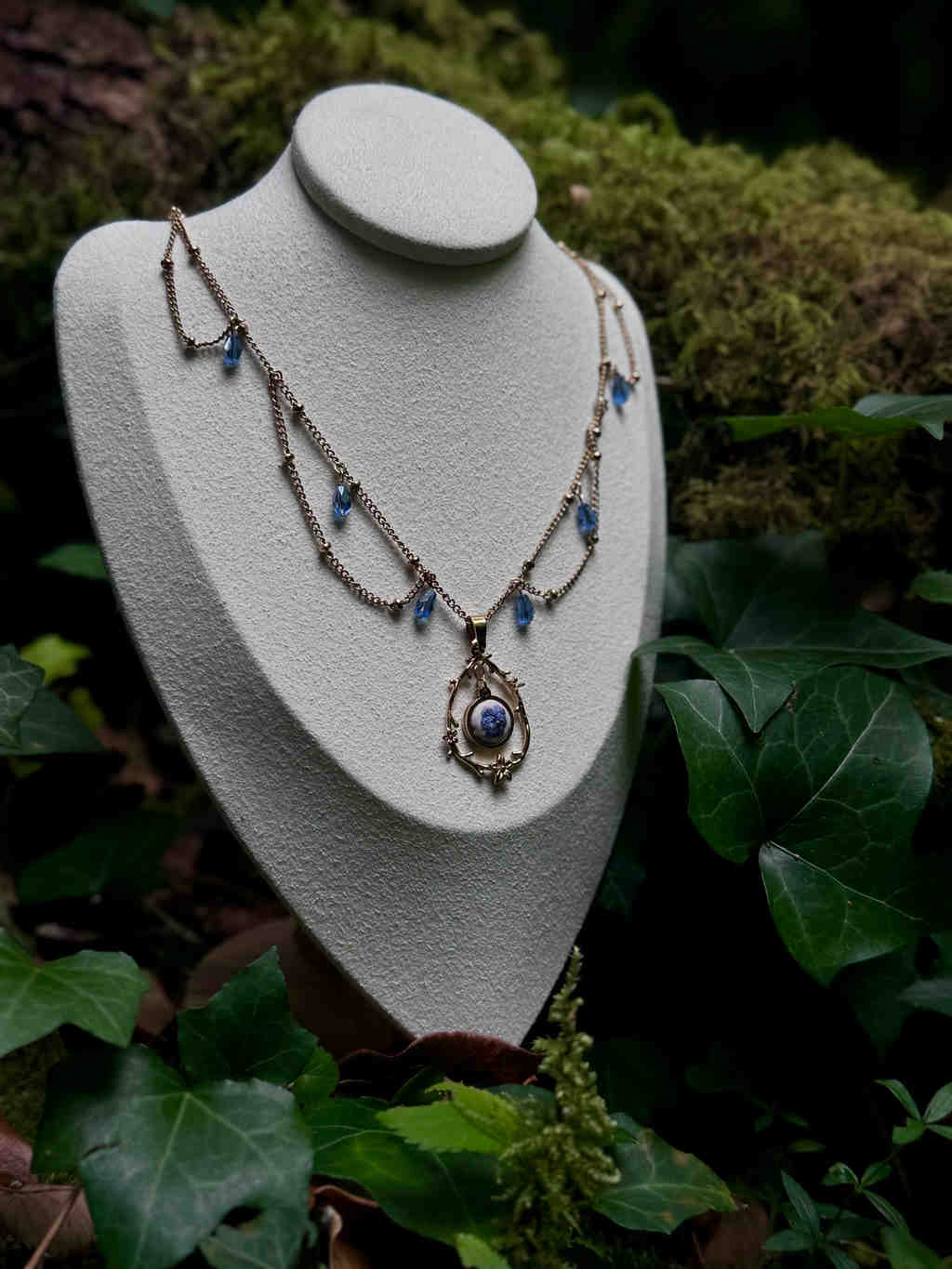 Cette image présente un collier délicat et orné, exposé sur un buste de présentation de bijoux de couleur claire, probablement en tissu ou en velours, qui contraste avec l'environnement naturel. Le collier est composé d'une chaîne fine de couleur dorée ou laiton, avec plusieurs rangées qui descendent en cascade. Il est agrémenté de petites perles ou cristaux bleus, de forme ovale ou en goutte, qui sont espacés le long des chaînes, ajoutant une touche de couleur et de brillance. Le pendentif central est la pièce maîtresse du collier. Il est de forme ovale, encadré par un motif ornemental détaillé, rappelant des volutes ou des feuilles stylisées, également de couleur dorée. À l'intérieur du pendentif, on peut distinguer une petite image ou un cabochon, qui semble représenter un paysage miniature ou un motif abstrait dans des tons de bleu et de blanc, évoquant peut-être un ciel nuageux ou une scène aquatique. Le buste de présentation est placé dans un cadre naturel, suggérant un environnement extérieur ou un décor inspiré de la nature. L'arrière-plan est flou, mais on peut distinguer des feuilles vertes foncées, du lierre et de la mousse, créant une ambiance forestière ou de jardin. La lumière semble douce et diffuse, mettant en valeur les détails du collier sans créer de reflets trop intenses. L'ensemble de l'image dégage une atmosphère de mystère, d'élégance et de connexion avec la nature, soulignant la beauté artisanale du bijou.