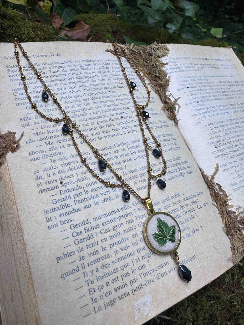 This image features a close-up, high-angle shot of a vintage-style necklace resting on an open book. The book, with aged, yellowed pages and French text, appears to be quite old, possibly a novel or a collection of stories. The necklace is made of a gold-toned chain, adorned with small, dark beads (possibly black or dark blue) interspersed along its length. The focal point of the necklace is a circular pendant with a gold-toned frame. Inside the frame, there's a white, fabric-like background on which a vibrant green ivy leaf is embroidered. Below the circular pendant, a single, teardrop-shaped dark bead hangs, matching the beads on the chain. The book is open to a page with dense text, and its spine is visible on the left side of the image. The pages are slightly crinkled and show signs of age. Around the book, especially on the right side and in the background, there are hints of a natural, mossy or earthy surface, suggesting the book and necklace are placed outdoors or in a setting with natural elements. Some dried, brown plant material is also scattered on and around the book, adding to the rustic and vintage aesthetic. The lighting in the image is soft, highlighting the details of the necklace and the texture of the book pages. The overall impression is one of charm, nostalgia, and a connection to nature.