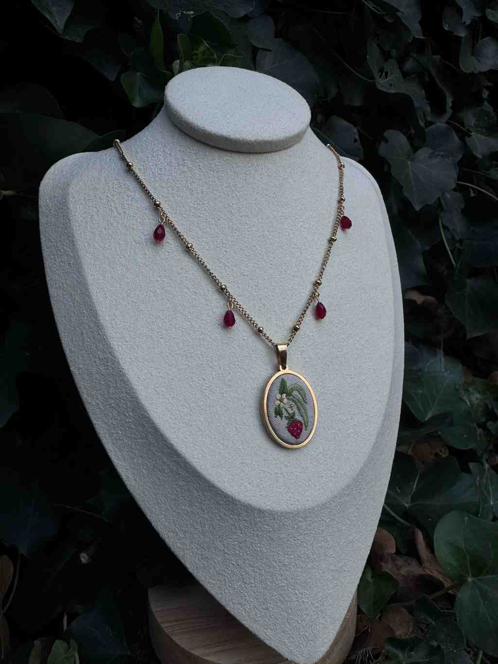 L'image présente un collier délicat et élégant, mis en valeur sur un buste de présentation de couleur crème ou beige clair. Le buste est placé devant un arrière-plan flou de feuillage vert foncé, probablement du lierre, ce qui crée un contraste naturel et met l'accent sur le bijou. Le collier est composé d'une fine chaîne dorée, parsemée de petites perles dorées à intervalles réguliers. Plusieurs petites perles rouges, de forme ovale ou en goutte, sont suspendues à la chaîne, ajoutant une touche de couleur et de raffinement. Le pendentif central est de forme ovale, encadré d'un fin liseré doré. À l'intérieur de l'ovale, on peut voir une broderie miniature ou une illustration détaillée. Le motif représente une petite fraise rouge vif avec ses feuilles vertes et des fleurs blanches délicates, le tout sur un fond clair. La finesse des détails de la broderie est remarquable, suggérant un travail artisanal minutieux. L'éclairage semble naturel, mettant en évidence les reflets dorés de la chaîne et du pendentif, ainsi que la texture du buste de présentation. L'ensemble dégage une impression de douceur, de féminité et de sophistication, évoquant la nature et l'artisanat.