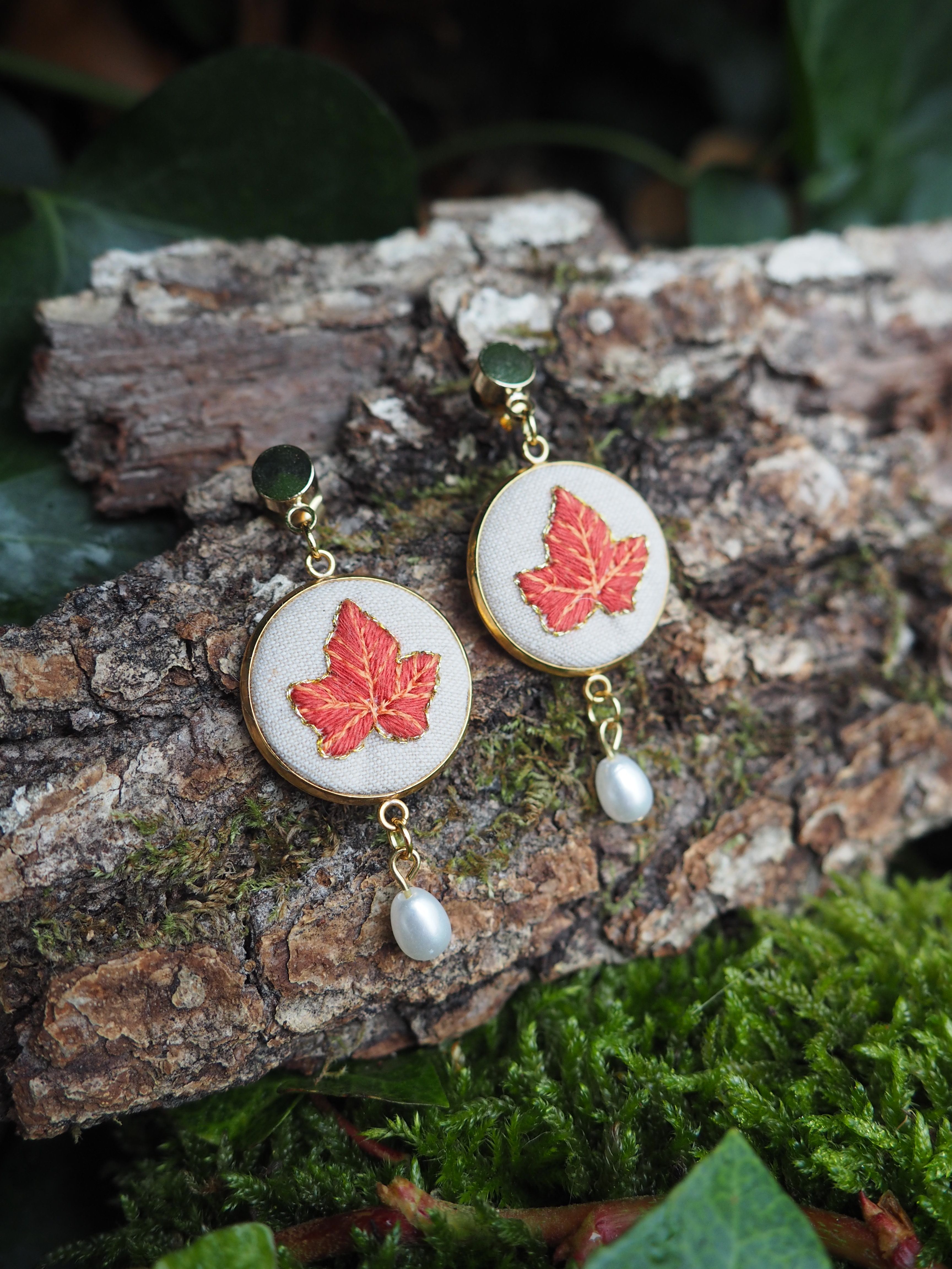 This close-up shot features a pair of elegant dangle earrings, each showcasing a beautifully embroidered red maple leaf on a circular, off-white fabric base. The embroidery is detailed, capturing the veins and shape of the leaf with vibrant red and subtle gold threads. The circular fabric is encased in a thin, golden metal frame, which also forms the stud and the dangle components of the earrings. Each earring is adorned with a small, lustrous white pearl that dangles gracefully from the bottom of the circular design, adding a touch of sophistication. The golden metal components appear polished and reflective. The earrings are artfully displayed on a piece of rough, textured tree bark, which is partially covered in patches of vibrant green moss. In the background, out of focus, are hints of dark green foliage, suggesting a natural, outdoor setting. The lighting is soft, highlighting the intricate details of the earrings and creating a gentle contrast with the rustic backdrop.