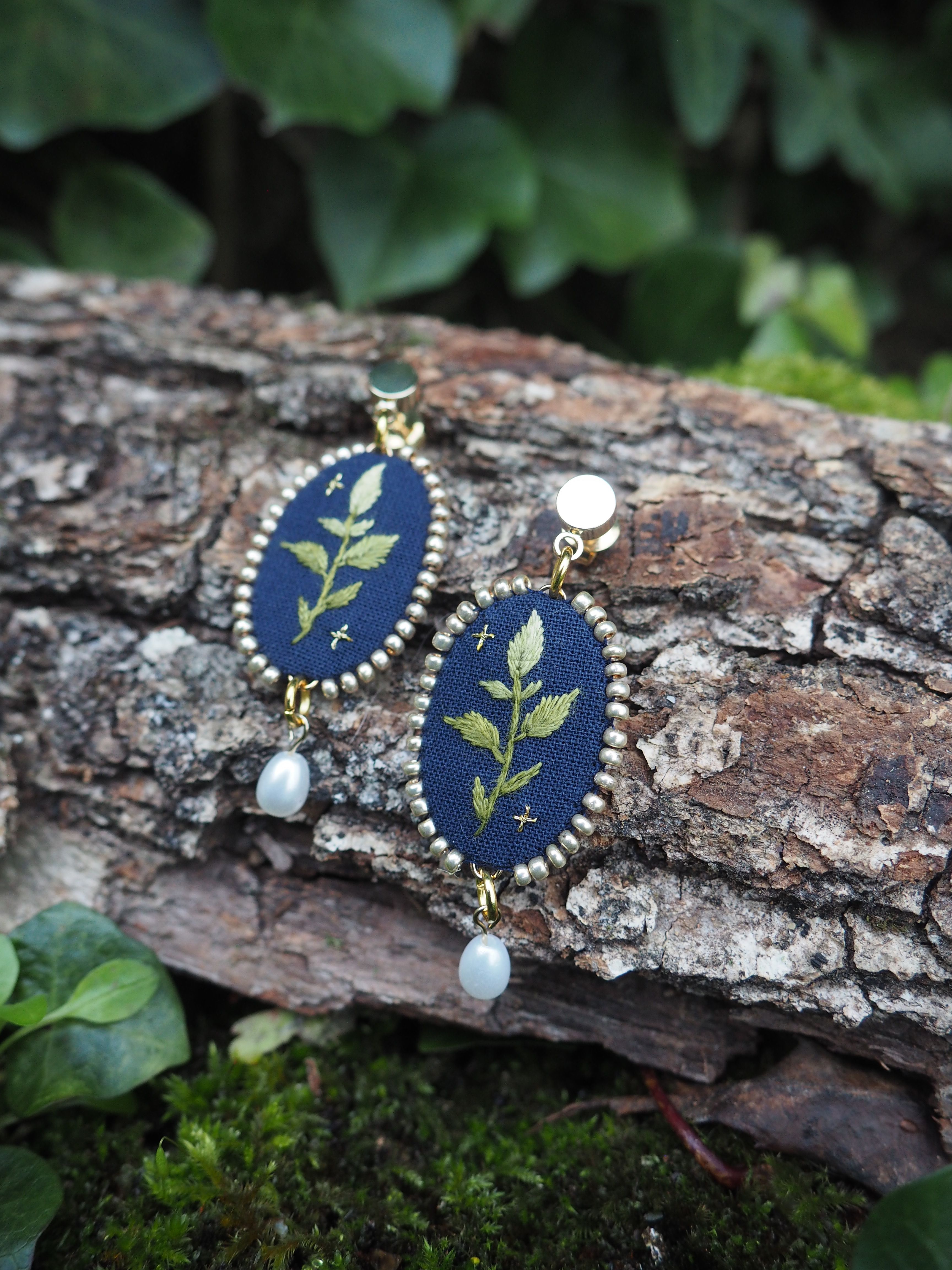 L'image montre une paire de boucles d'oreilles artisanales posées sur un morceau d'écorce d'arbre. Les boucles d'oreilles sont ovales avec un fond bleu foncé sur lequel est brodé un motif de feuille ou de branche en vert clair. Le contour des ovales semble être en métal doré, donnant un aspect élégant à l'ensemble. Chaque boucle d'oreille est ornée d'une petite perle blanche suspendue en bas, ajoutant une touche délicate. Le contraste entre le bleu profond du fond et le vert tendre de la broderie crée un effet visuel très attrayant. L'arrière-plan montre l'écorce rugueuse et texturée sur laquelle les boucles d'oreilles sont posées, ainsi que quelques feuilles vertes floues en arrière-plan, suggérant un cadre naturel en extérieur. Cette mise en scène met en valeur l'aspect artisanal et naturel des bijoux. Ces boucles d'oreilles semblent être un bel exemple d'artisanat mêlant broderie et joaillerie, avec une inspiration clairement tirée de la nature.