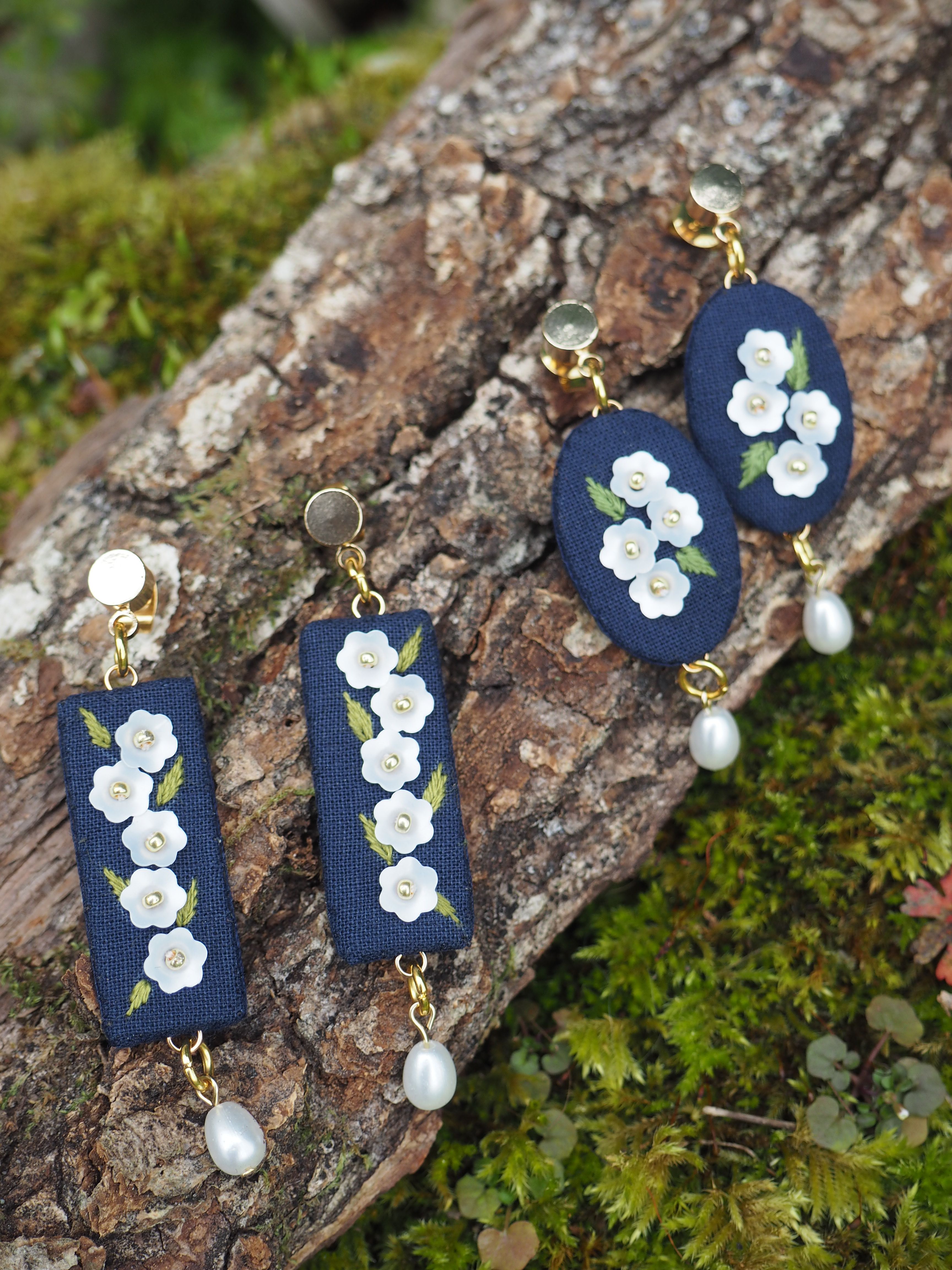 L'image montre des bijoux délicats posés sur un morceau d'écorce d'arbre couvert de mousse. Il s'agit de trois paires de boucles d'oreilles artisanales. Les boucles d'oreilles sont composées de petits rectangles ou ovales en tissu bleu marine, sur lesquels sont brodés des motifs floraux blancs qui ressemblent à de petites fleurs. Chaque paire est légèrement différente dans sa forme, mais toutes partagent le même thème floral délicat. Au bas de chaque boucle d'oreille pend une petite perle blanche, donnant une touche d'élégance supplémentaire. Les attaches des boucles d'oreilles semblent être en métal doré. Le contraste entre les bijoux raffinés et le fond naturel rugueux de l'écorce et de la mousse verte crée une image très esthétique. Cela met en valeur la délicatesse et la beauté artisanale des boucles d'oreilles tout en les plaçant dans un contexte naturel et organique. Cette présentation suggère que ces bijoux pourraient être des créations artisanales ou inspirées par la nature, peut-être faits à la main avec des matériaux naturels ou recyclés.