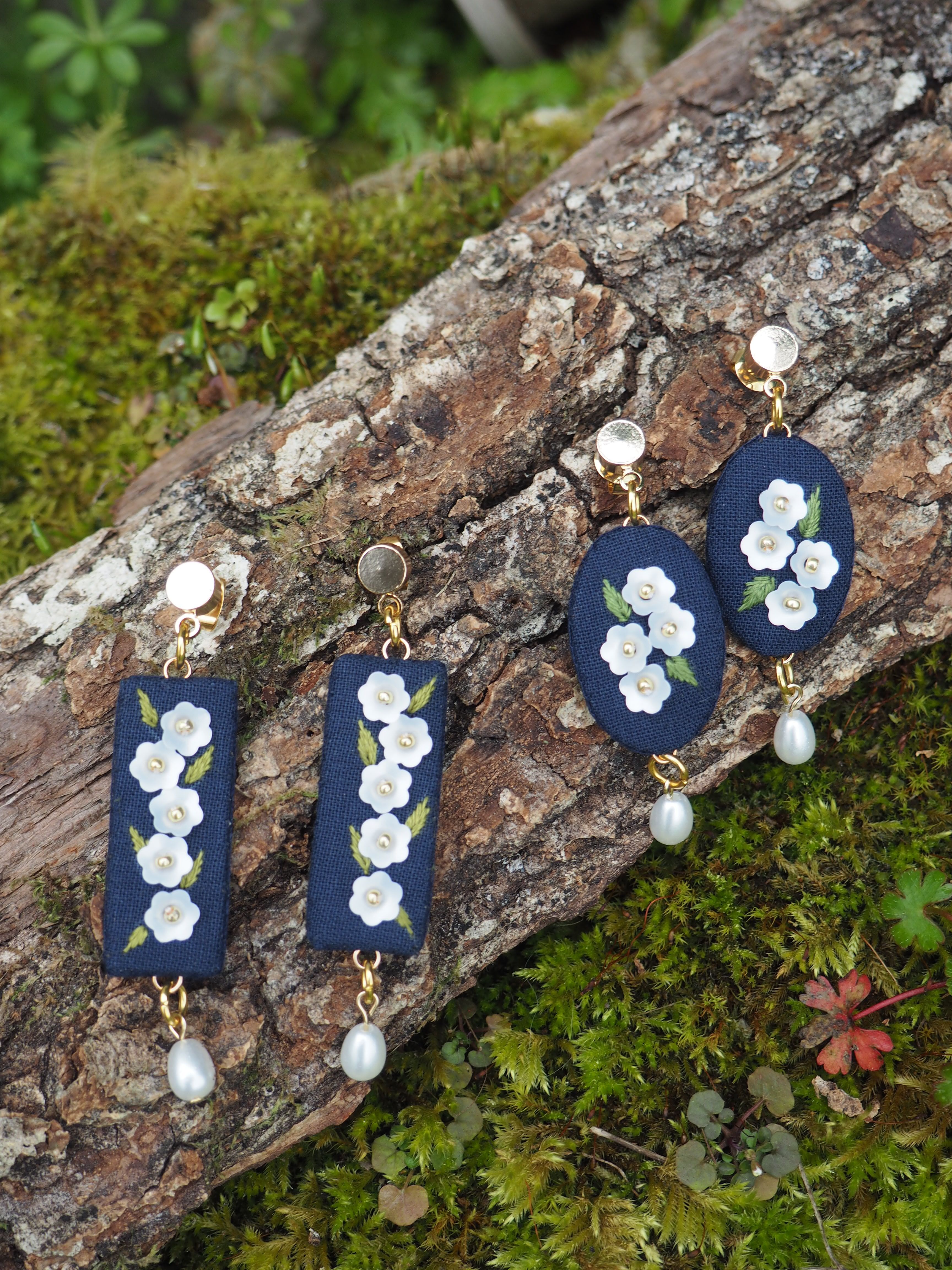L'image montre des bijoux délicats posés sur un morceau de bois couvert de mousse dans un environnement naturel. Les bijoux semblent être des boucles d'oreilles faites à la main. Il y a deux paires : une paire de pendentifs rectangulaires et une paire de pendentifs ronds. Les boucles d'oreilles ont une base bleu foncé, probablement en tissu ou en cuir, sur laquelle sont appliquées de petites fleurs blanches, qui ressemblent à des fleurs de cerisier ou de pommier. Chaque paire est ornée d'une perle blanche suspendue au bas. Les bijoux reposent sur l'écorce rugueuse et texturée d'un tronc d'arbre ou d'une branche. Autour, on peut voir de la mousse verte luxuriante et quelques petites plantes, créant un cadre naturel et forestier pour ces délicates pièces de bijouterie. Cette mise en scène met en valeur le contraste entre la délicatesse des bijoux faits main et la robustesse de la nature environnante, suggérant peut-être une inspiration naturelle dans la conception de ces accessoires.